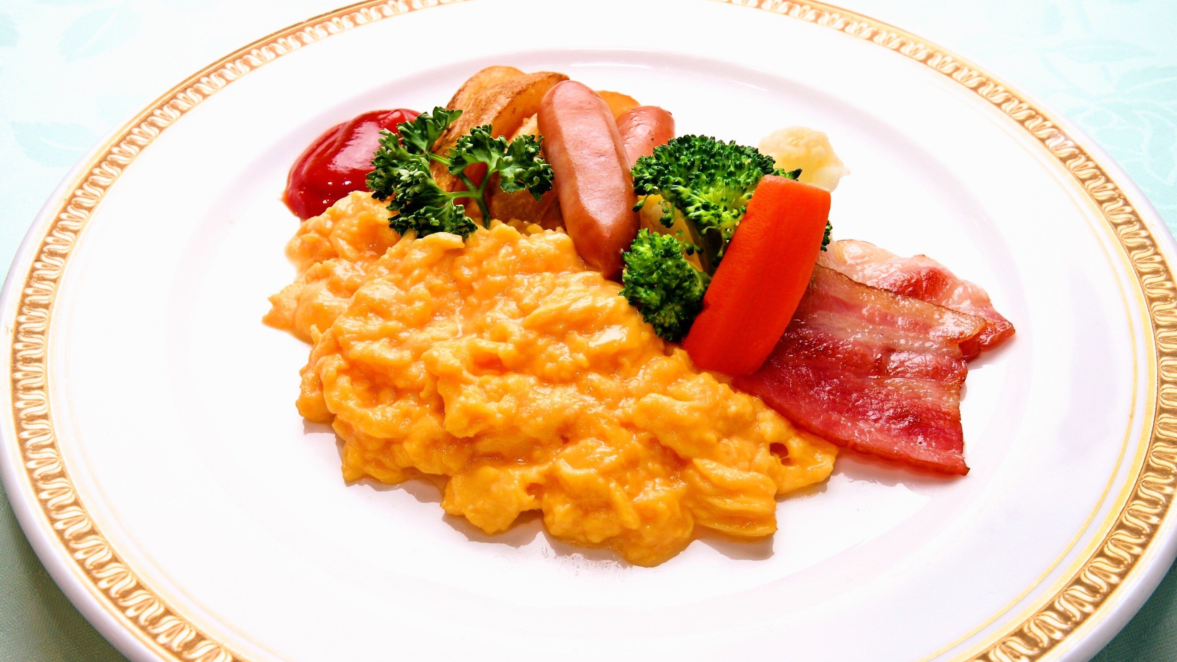 A plate of scrambled eggs, bacon, sausages, and vegetables on a white plate with a gold rim.