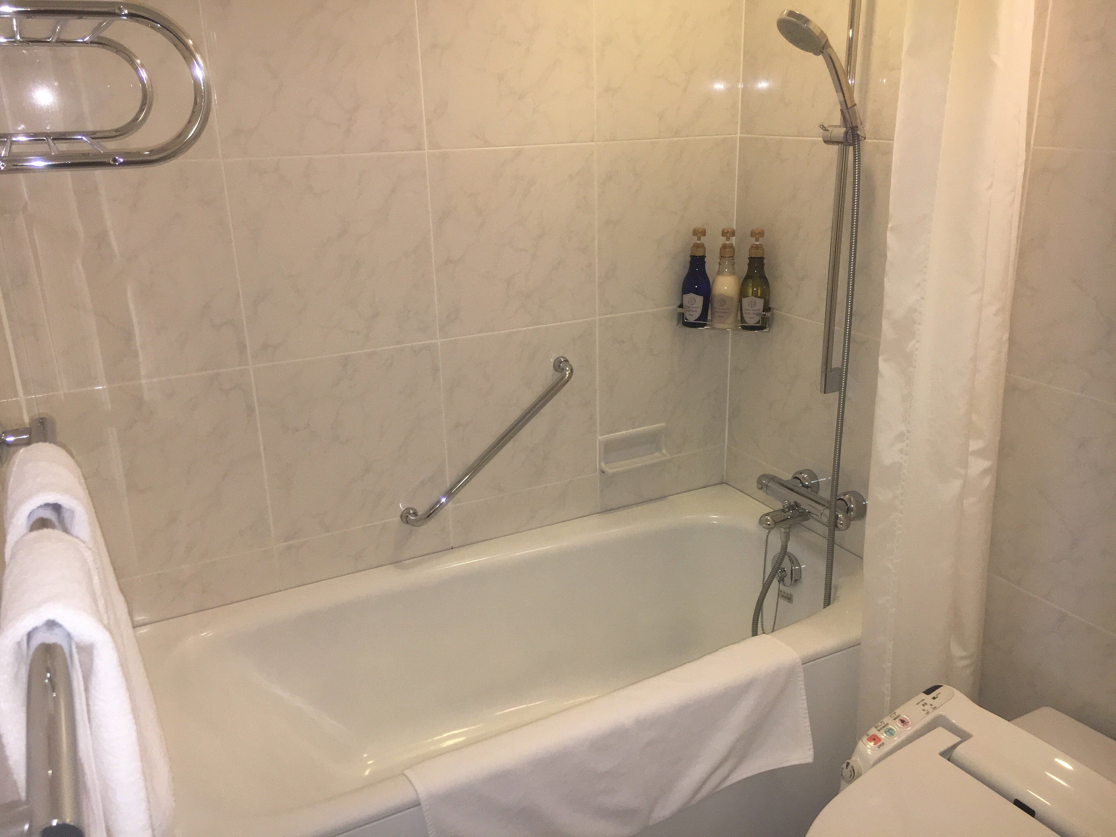 Un bagno moderno con vasca, doccia e mancorrente. Sulla mensola sopra la vasca ci sono delle bottiglie.