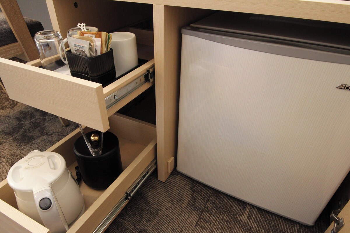 A compact kitchenette with a refrigerator, a coffee maker, a kettle, and a drawer containing coffee pods and a glass.