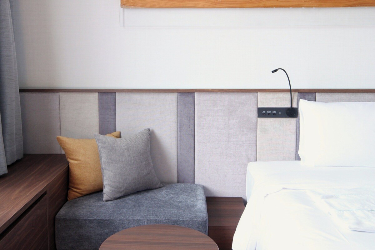 A hotel room with a bed, bedside table, and a seating area with cushions.