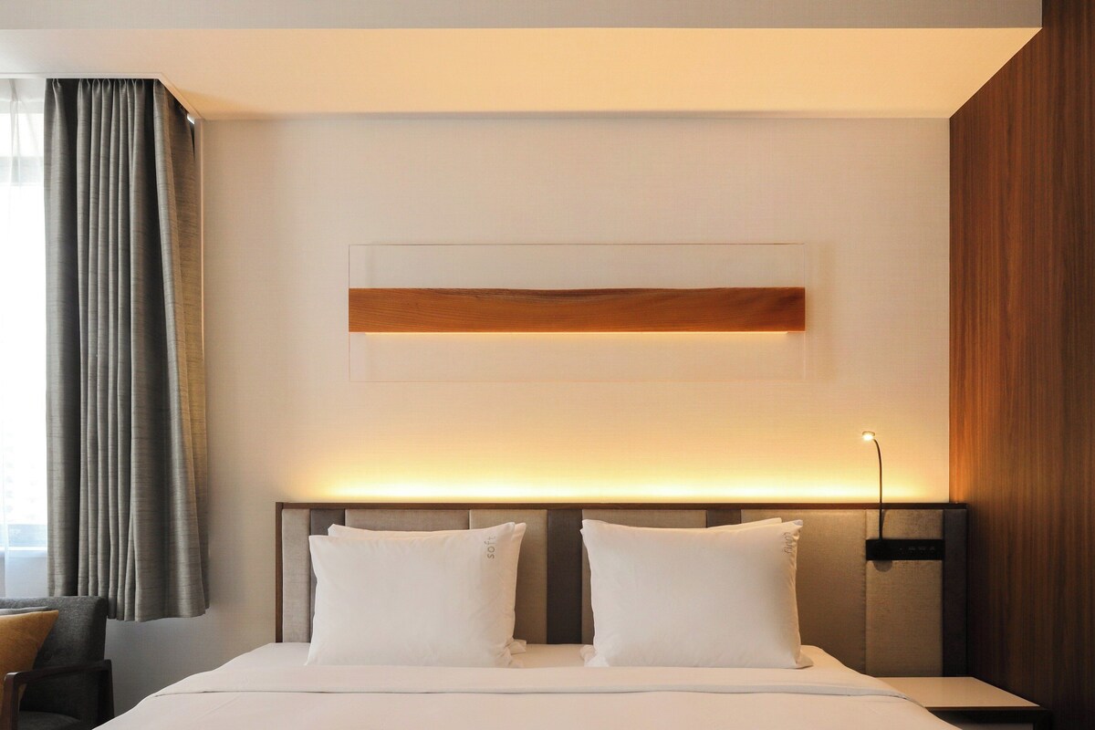A hotel room with a bed, two pillows, a bedside table, a lamp, and a large window with curtains.
