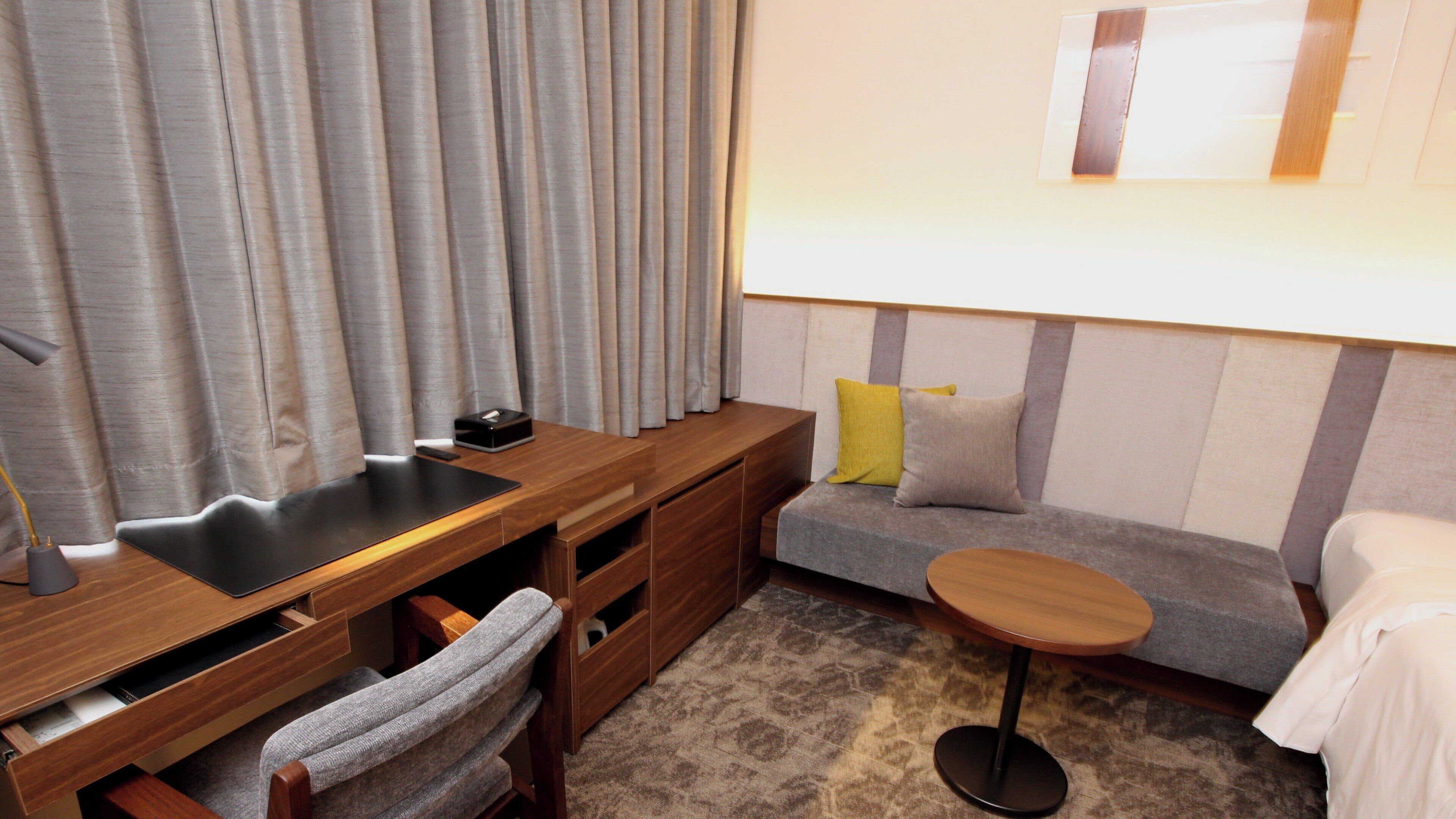 A modern hotel room with a wooden desk, a small round table, a sofa with cushions, and a large window with curtains.