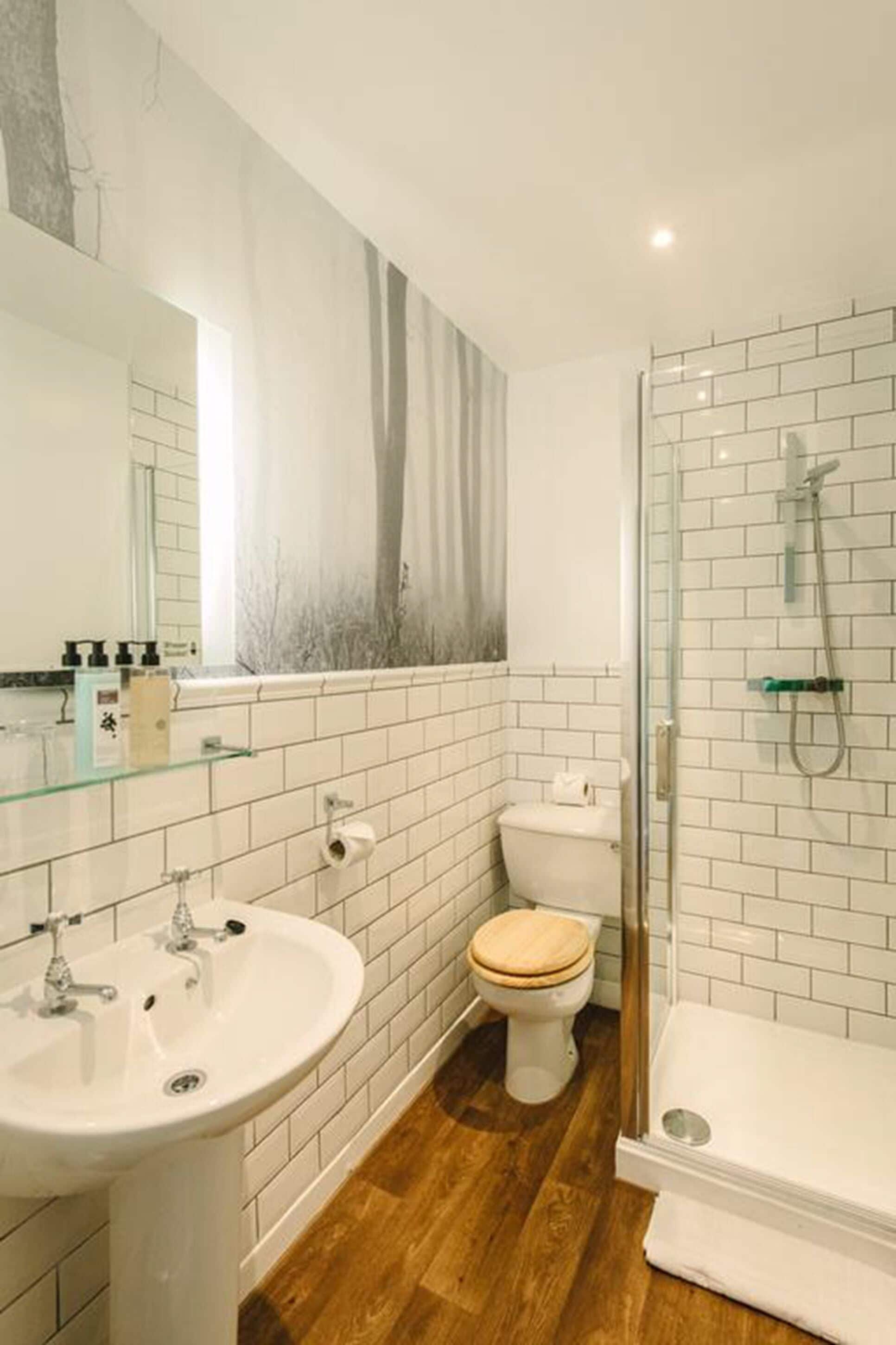 Un bagno moderno con pareti piastrellate di bianco, pavimento in legno, box doccia in vetro e uno specchio grande.