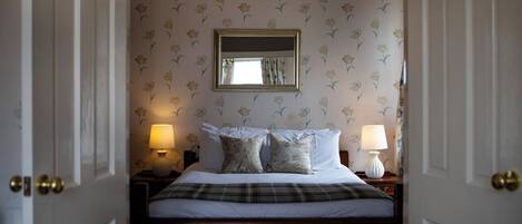 A bedroom with a bed, two bedside lamps, a mirror, and floral wallpaper.