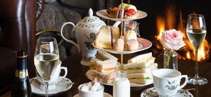 A table set with a teapot, teacups, a bottle of wine, a glass of wine, a rose, and a tiered tray of sandwiches and pastries. - The Boat Hotel (Boat of Garten)