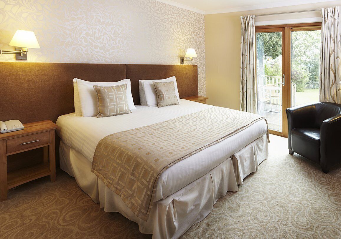 A hotel room with a large bed, two bedside tables, a nightstand, a chair, and a view of the outdoors through a glass door.