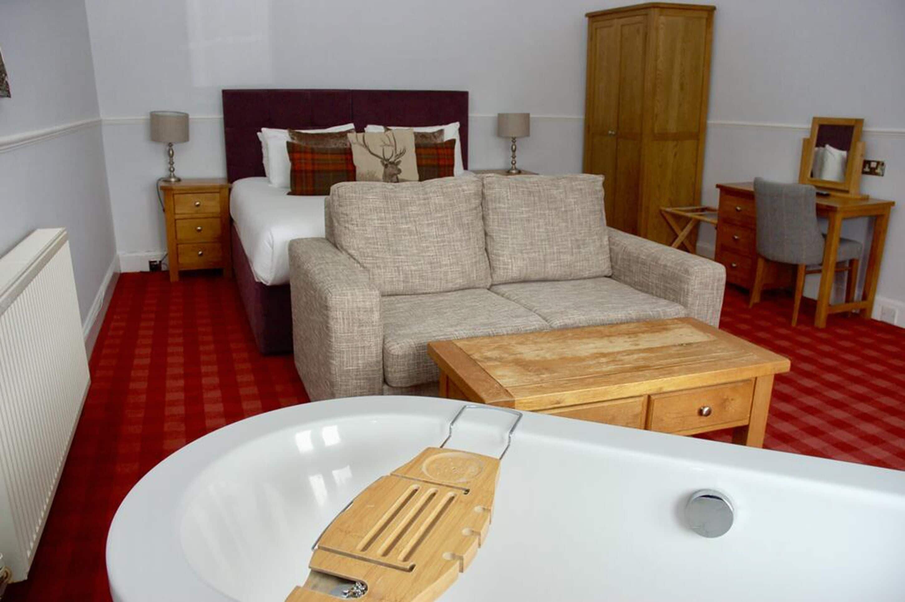 A bedroom with a bed, a sofa, a wooden table, a desk, and a large white bathtub.