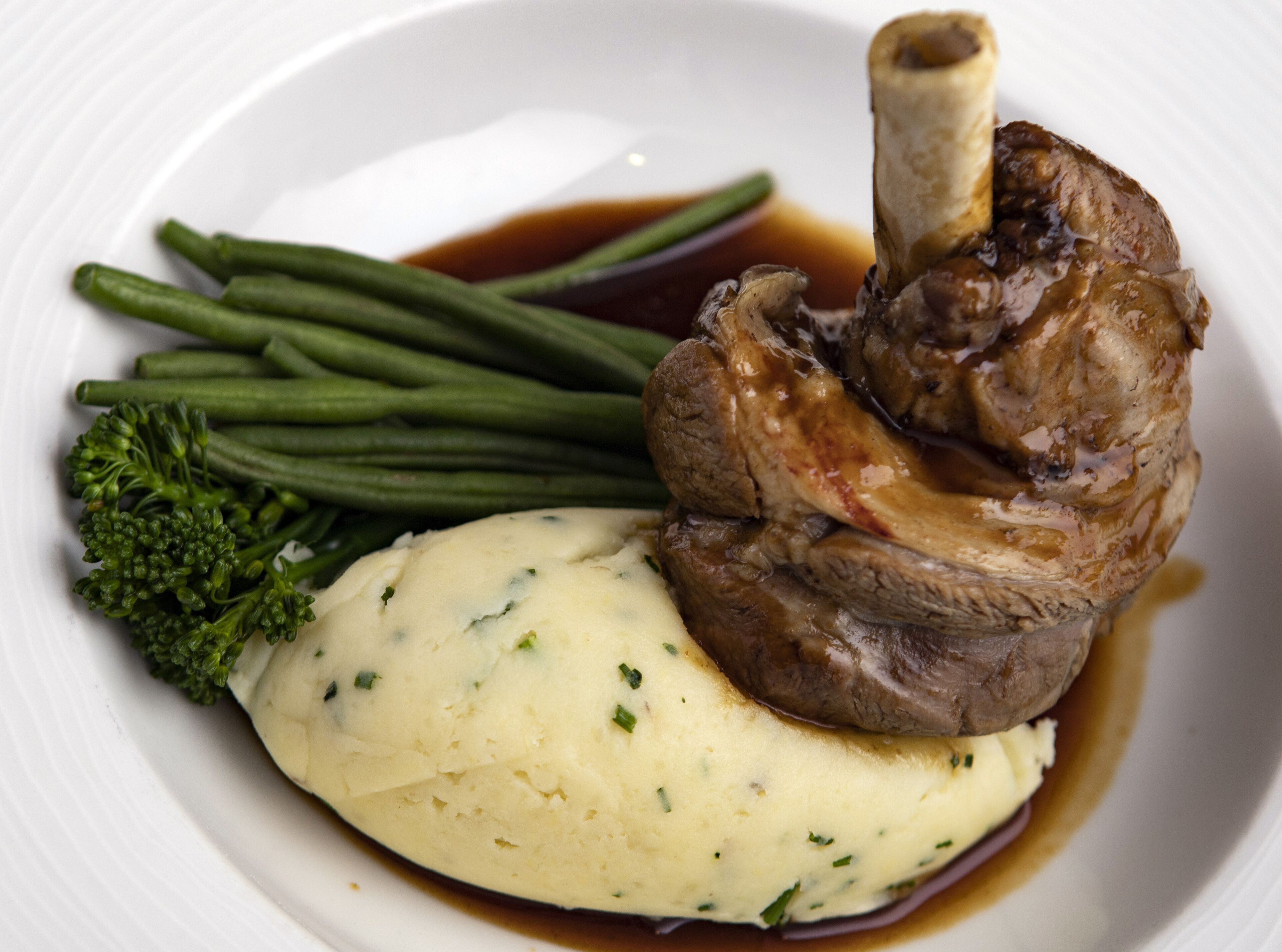 A plate with a lamb shank, green beans, and mashed potatoes.
