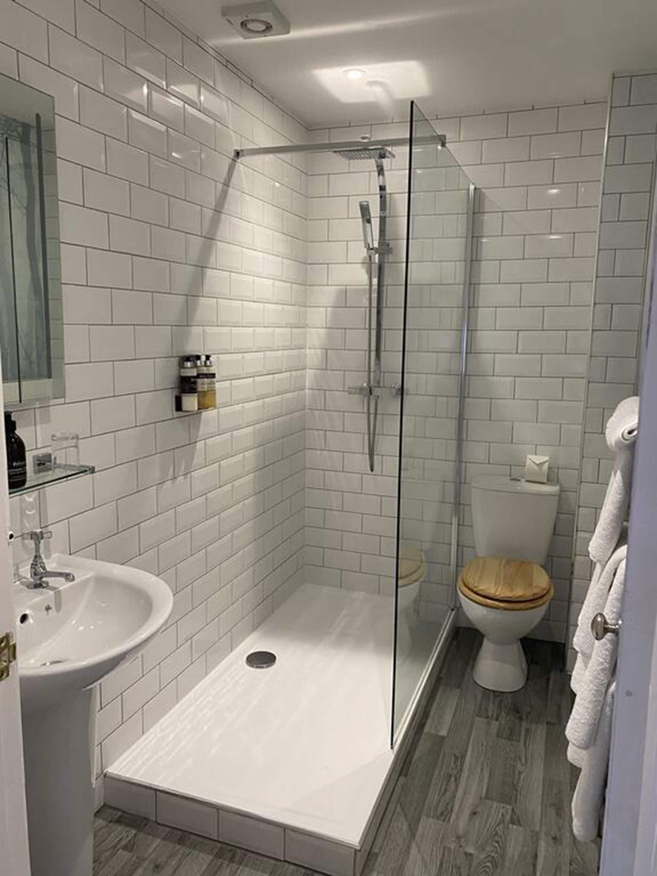 Ein modernes Badezimmer mit weißen Fliesen, einer Duschkabine, einem weißen Waschbecken und einer Toilette mit Holzsitz.