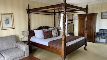 A four-poster bed with a plaid bedspread, a grey sofa, a wooden coffee table, and a plaid armchair in a room with striped wallpaper and a framed picture on the wall.