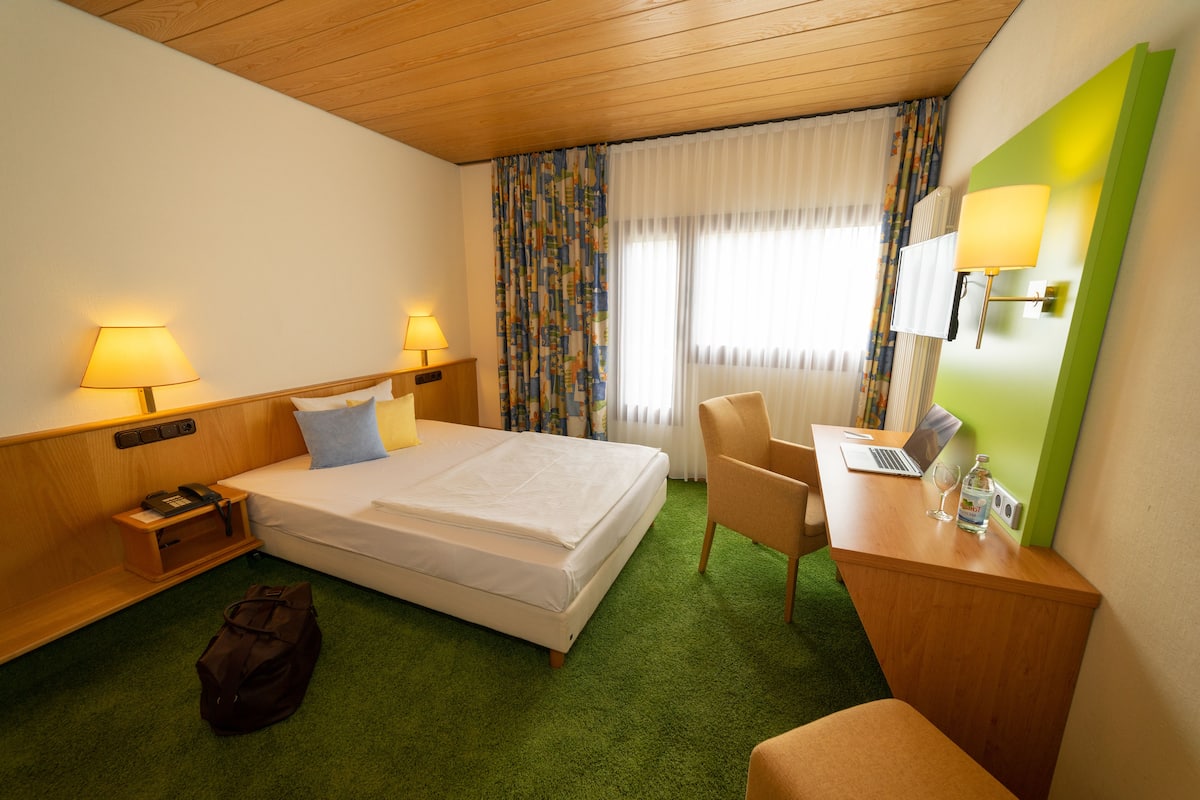 A hotel room with a bed, desk, chair, and a lamp.