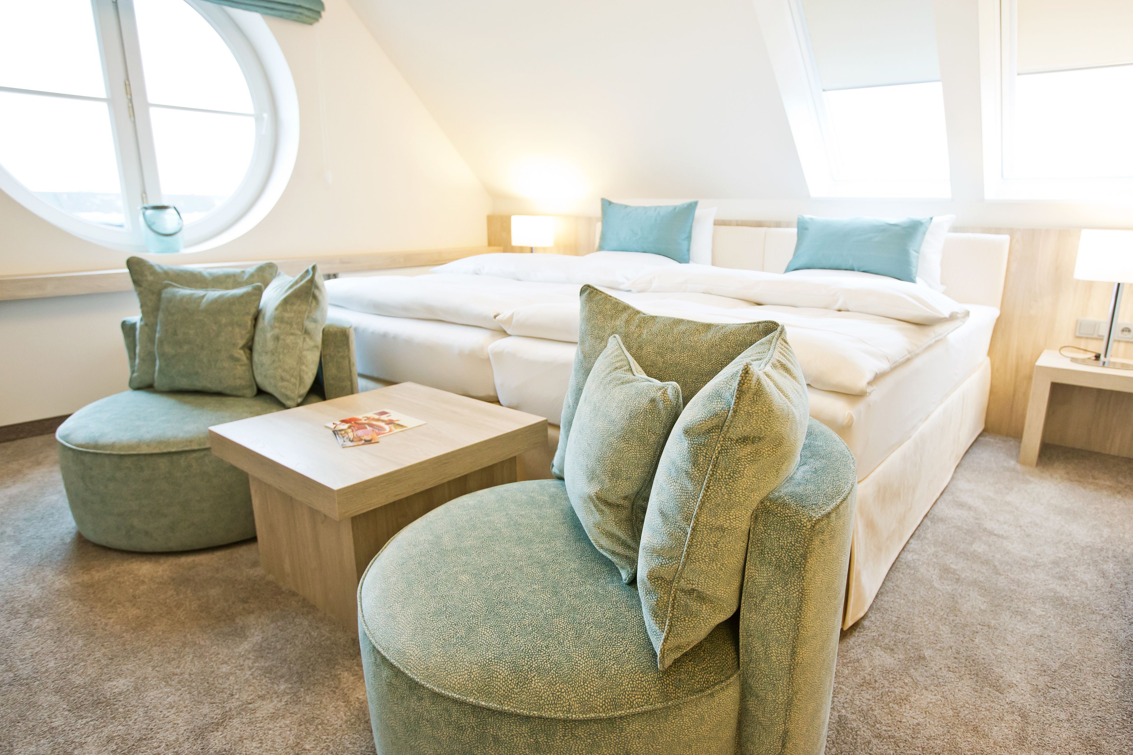 A modern hotel room with a bed, two green armchairs, a wooden coffee table, and a round window.