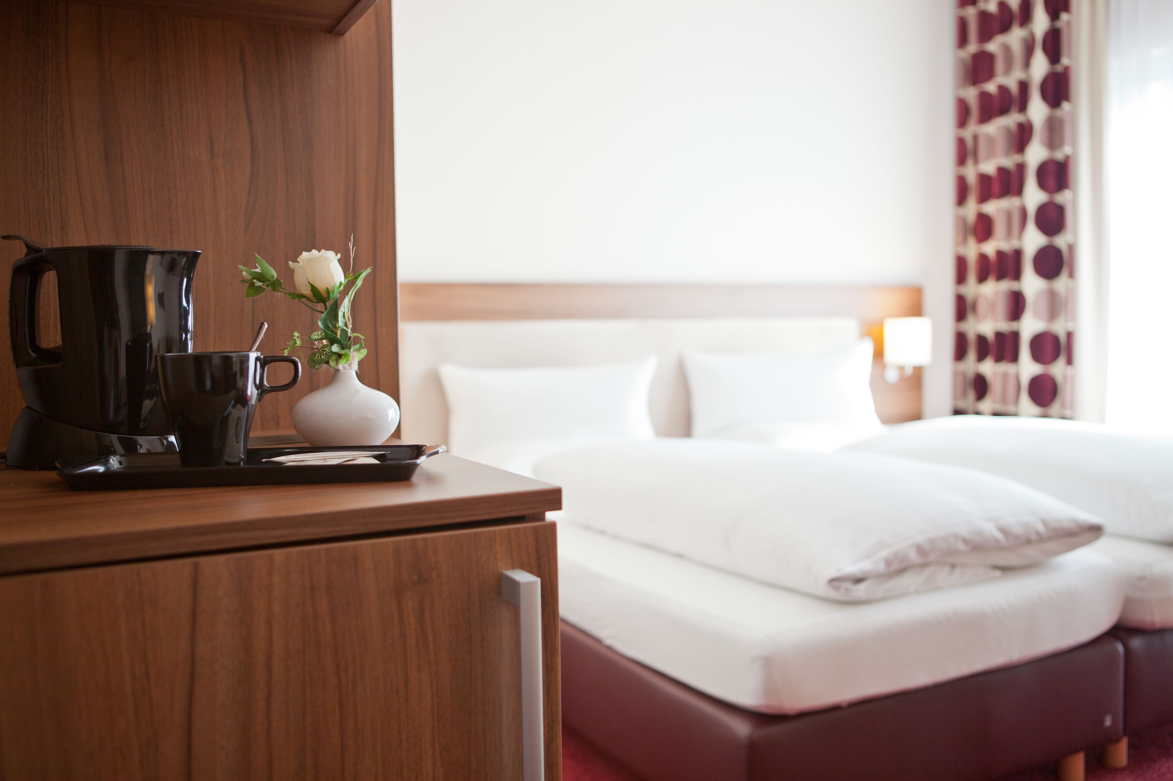 A hotel room with a wooden desk, a black coffee maker, a white vase with a flower, and a bed with white bedding.