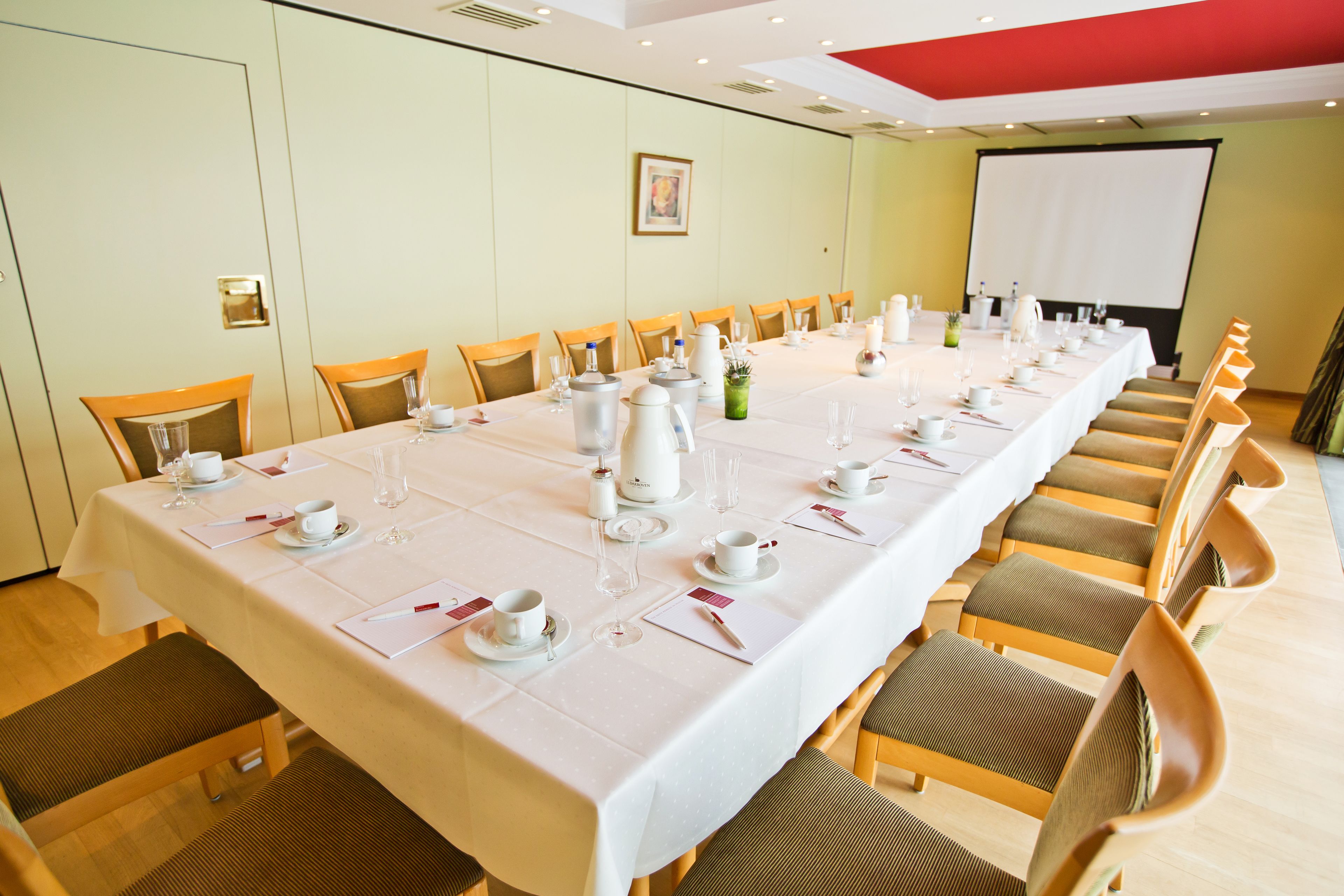Un salón de conferencias con una larga mesa dispuesta para una reunión, equipado con sillas, una pantalla de proyector y un mantel blanco.