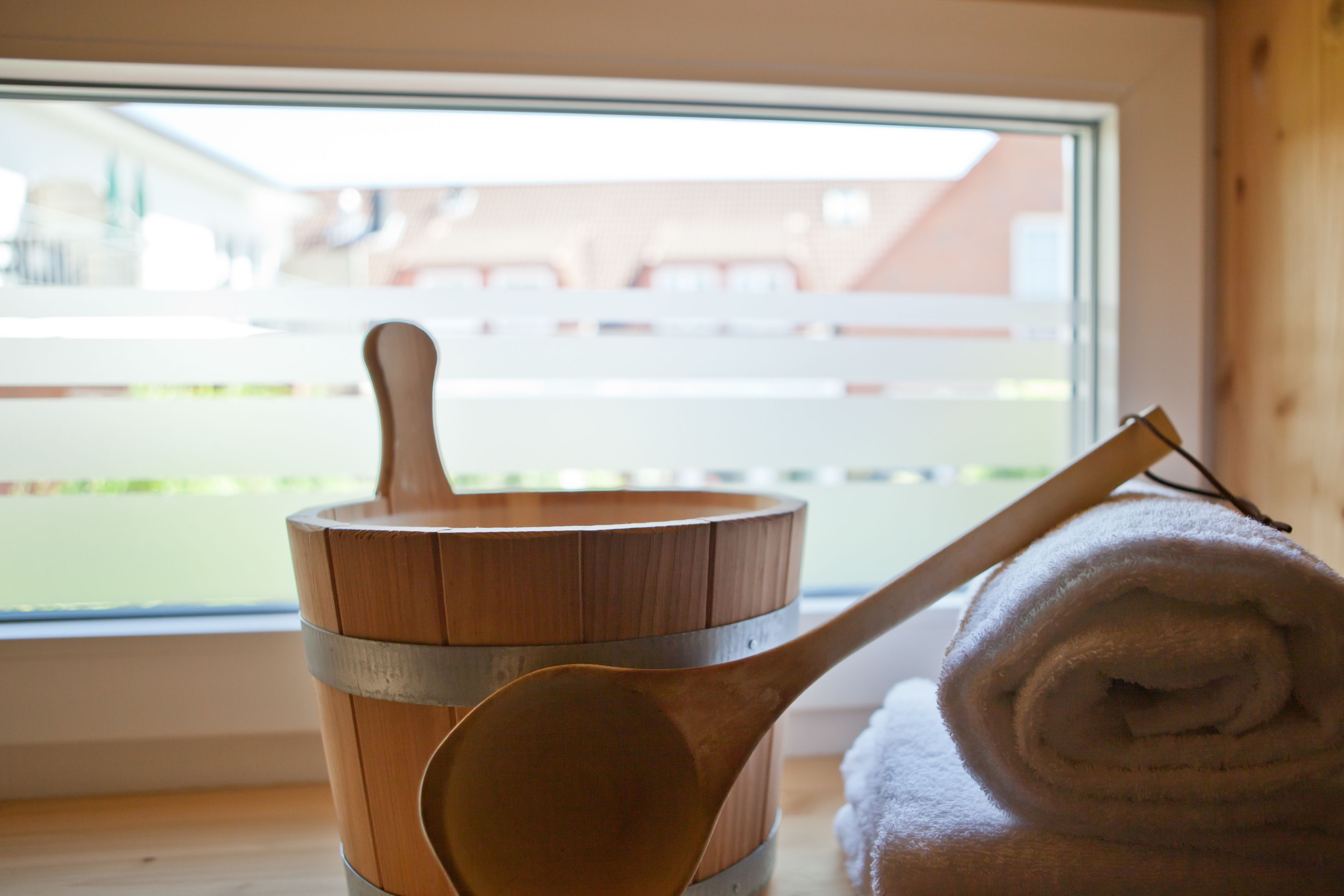 A wooden bucket with a wooden ladle, a rolled white towel, and a window with a view of a fence and buildings.