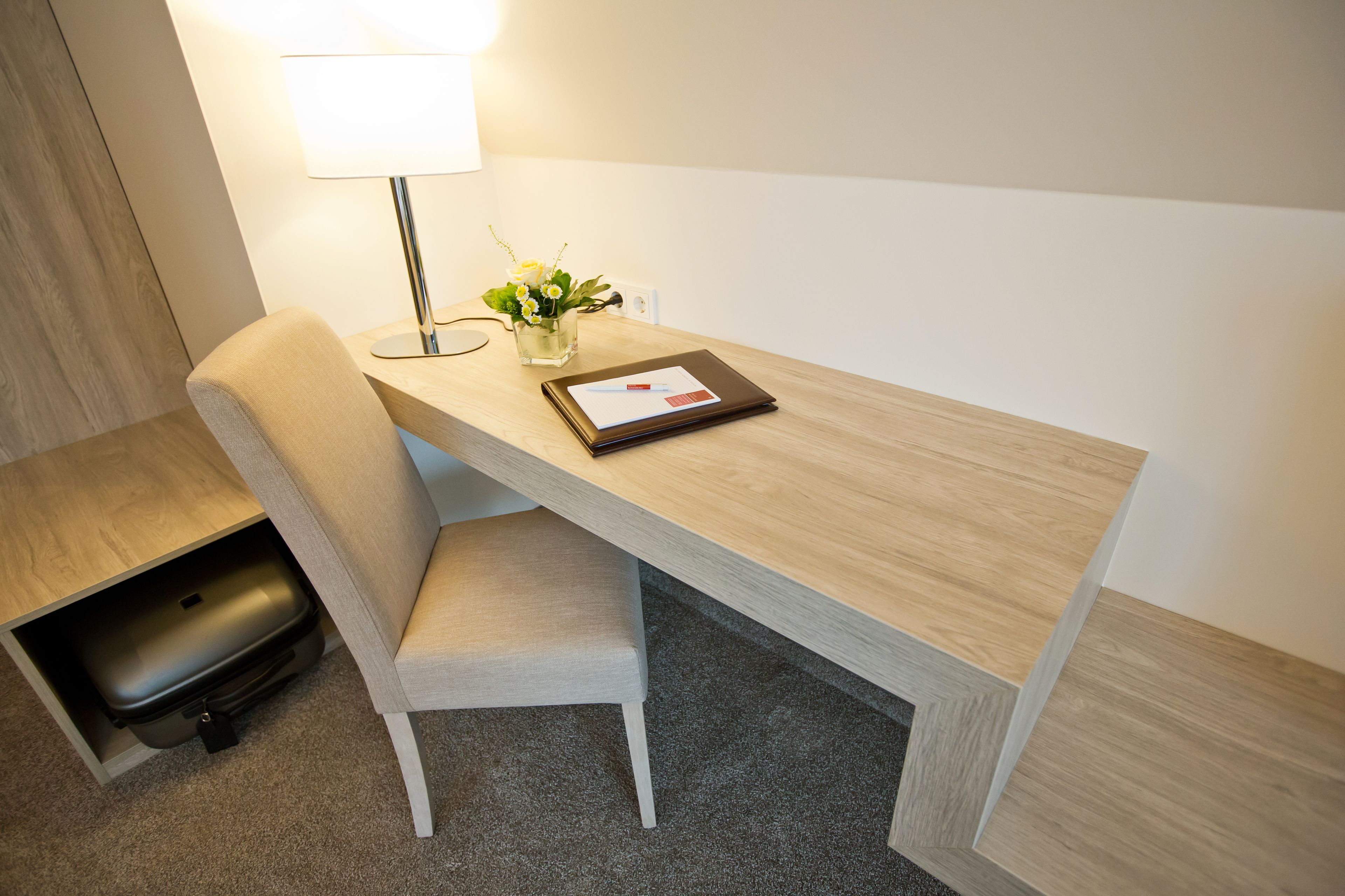 A hotel room with a wooden desk, a chair, a lamp, a small vase with flowers, and a tablet.