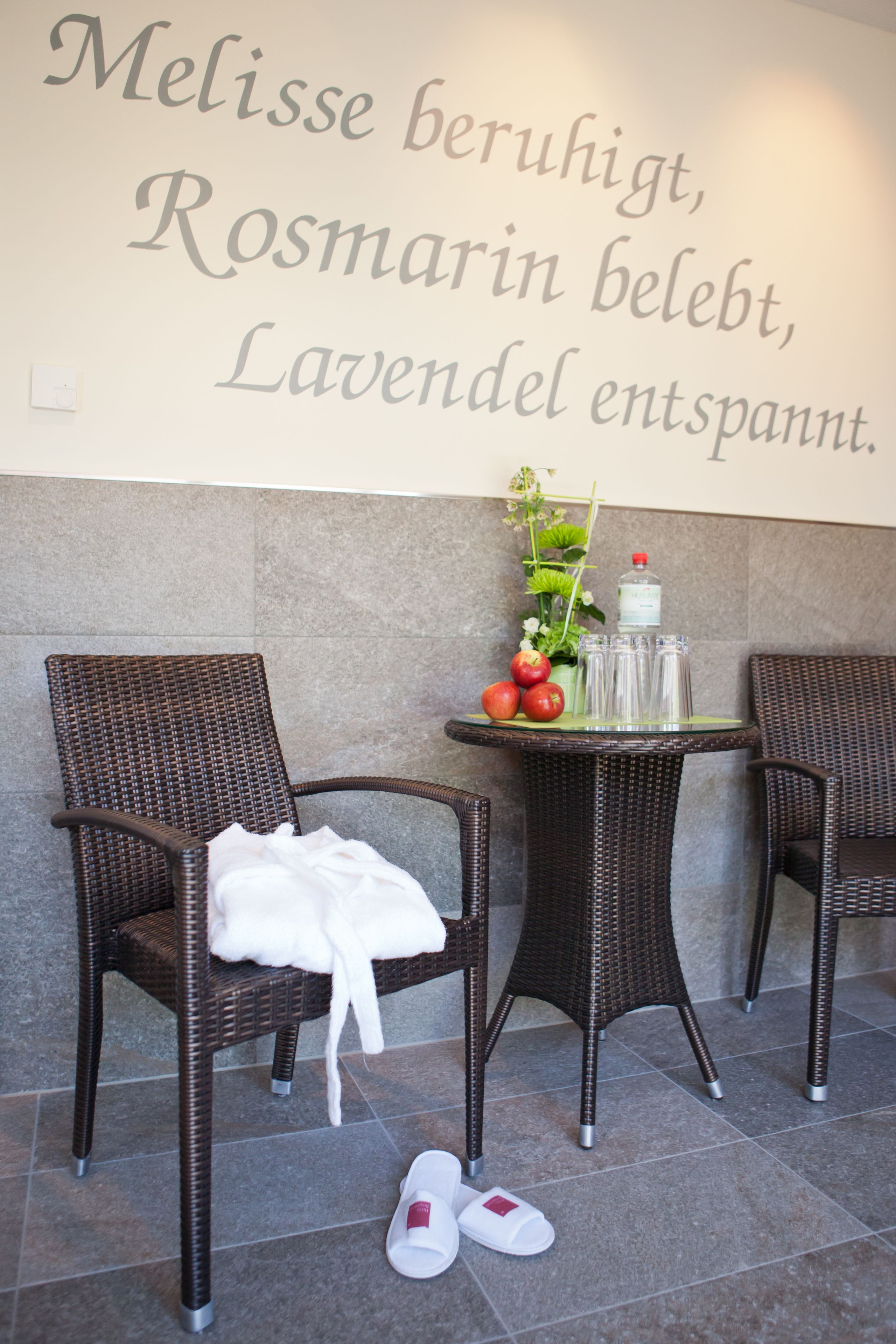 Eine Sitzecke mit Korbstühlen, einem kleinen Tisch mit Pflanze und Flasche, und einer Wand mit einem deutschen Text.