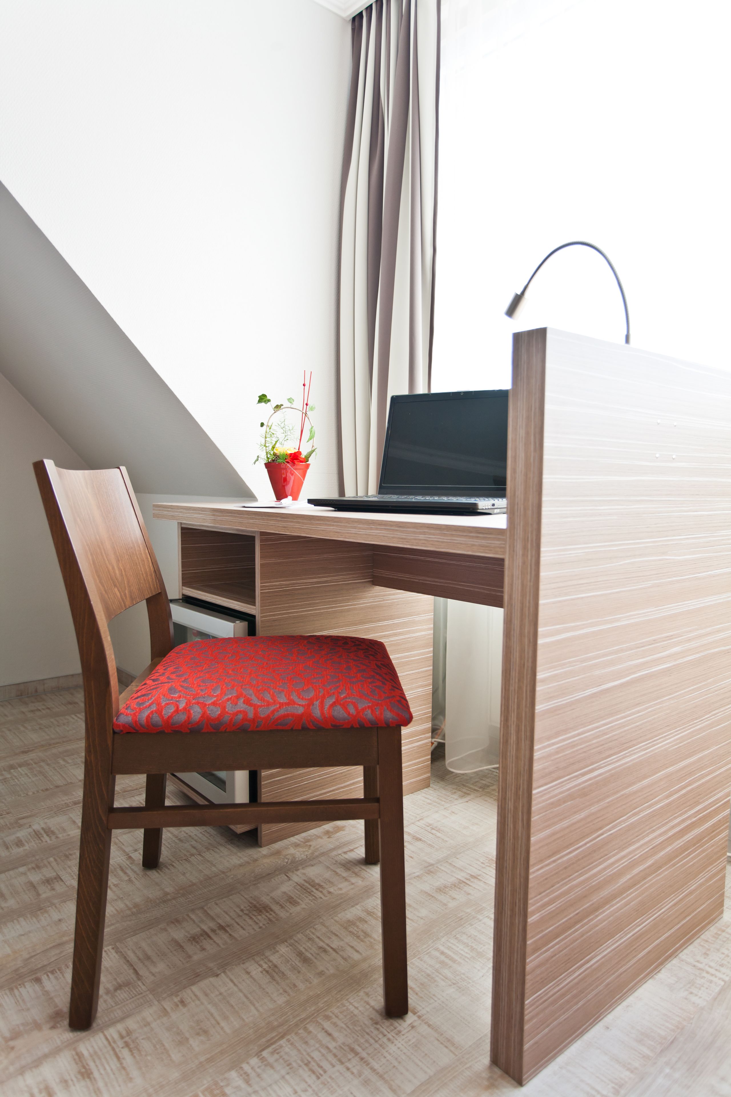 A modern workspace with a wooden desk, a laptop, a red cushioned chair, and a potted plant.