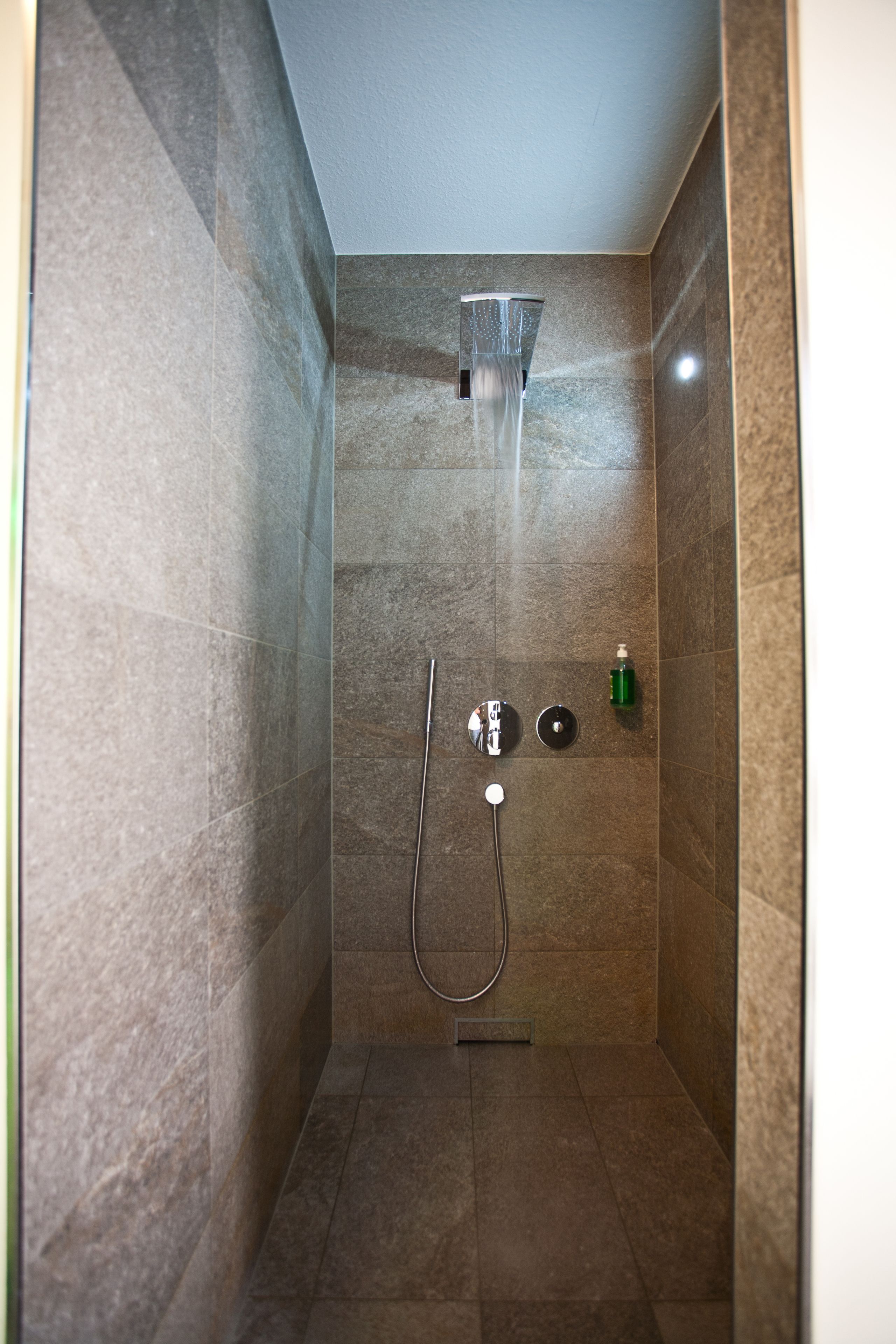 Een kleine badkamer met betegelde wanden, een handdouche, een kraan op de muur en een zeepdispenser.