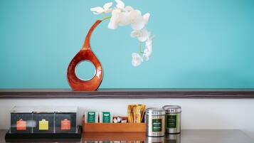 Un jarrón de madera con orquídeas blancas sobre una pared verde azulada, una presentación de bolsitas de té y dos recipientes metálicos sobre una superficie reflectante.