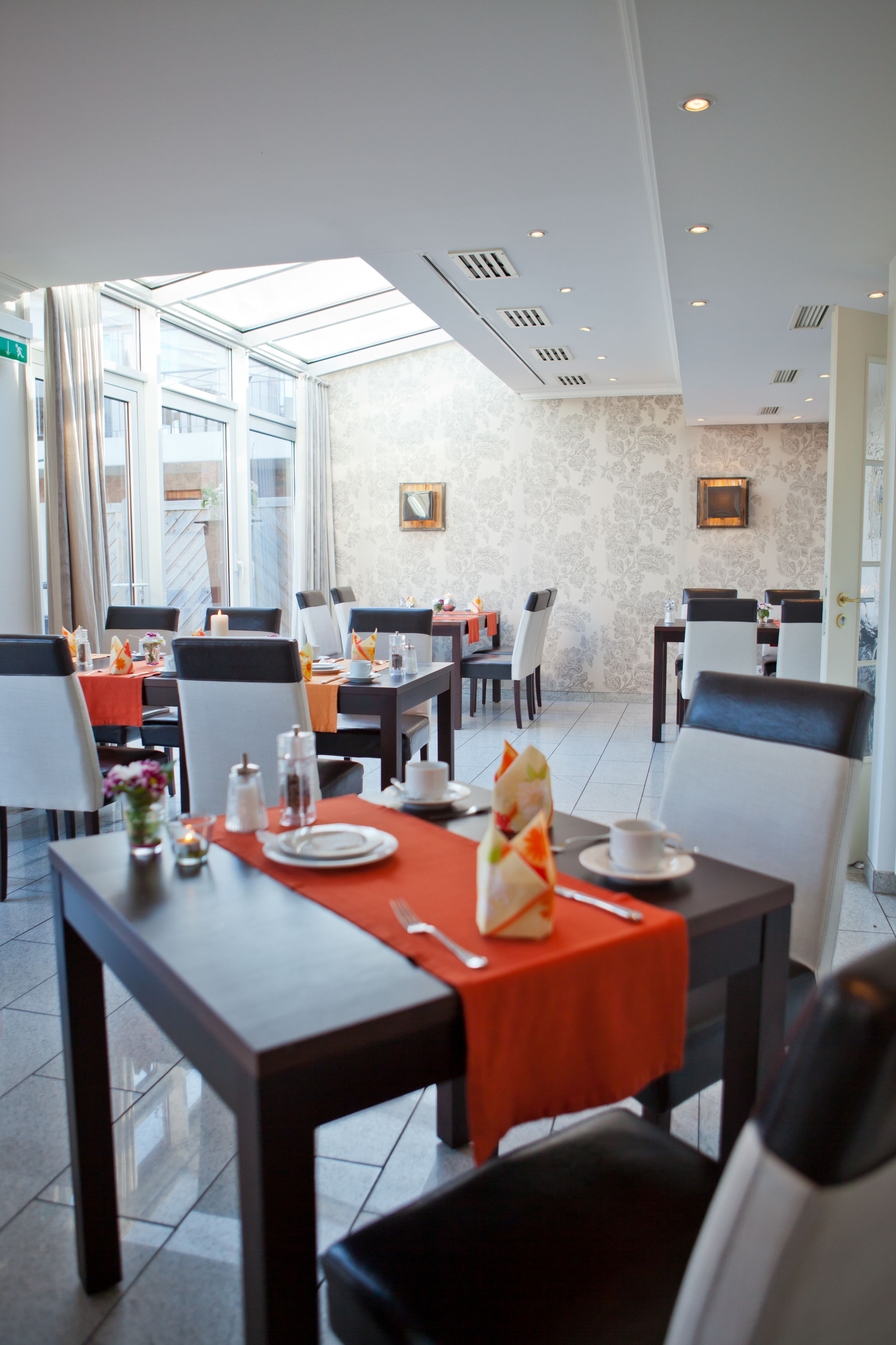A modern dining area with tables set for two, featuring a bright, naturally lit space with large windows and a high ceiling.