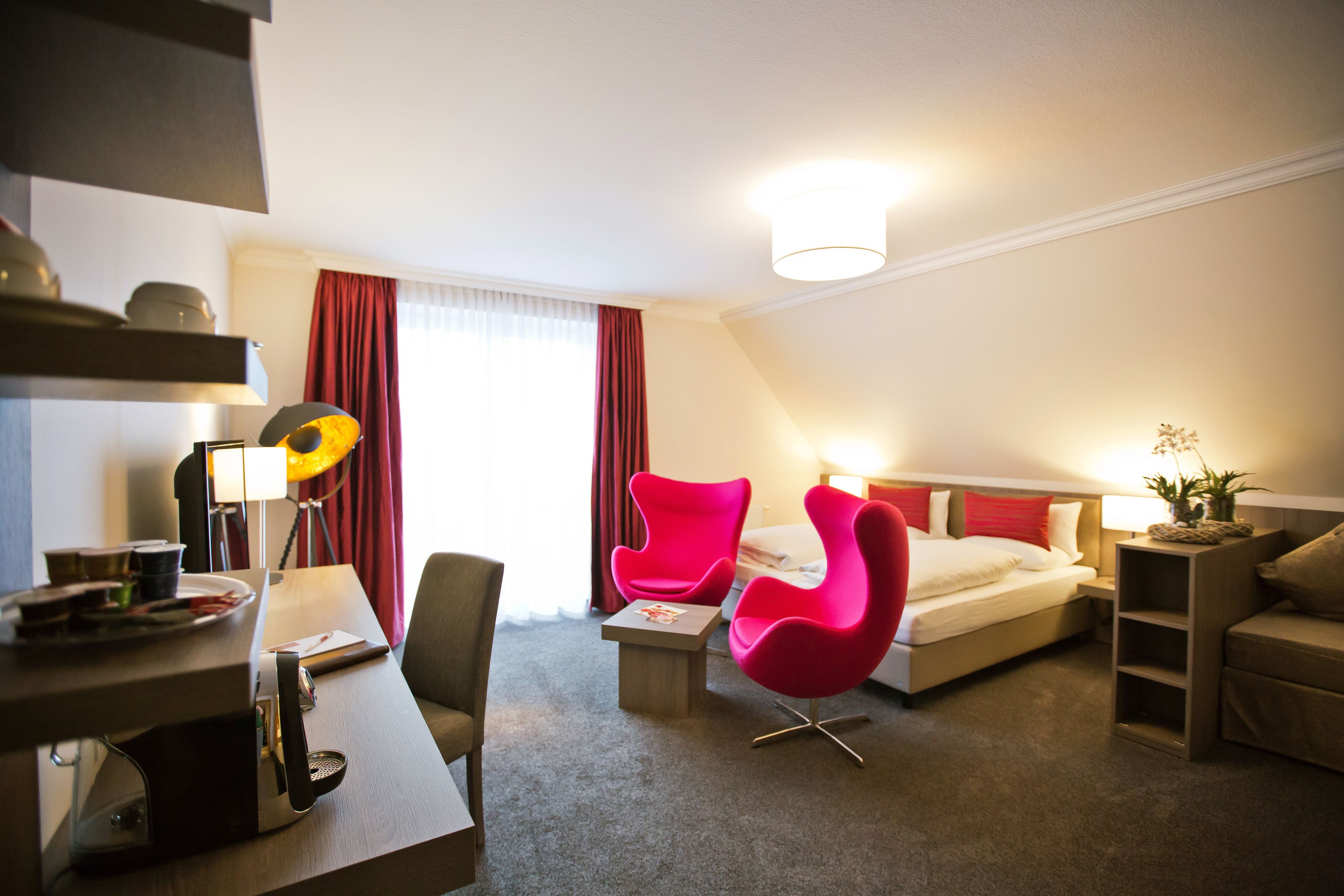 A hotel room with a bed, a desk with a lamp, a chair, and a small table.