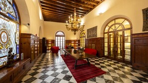 Um hall de hotel com piso xadrez, um candelabro, mobiliário de madeira e janelas de vitral.