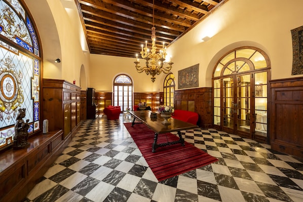 A hotel lobby with a checkered floor, a chandelier, wooden furniture, and stained glass windows.