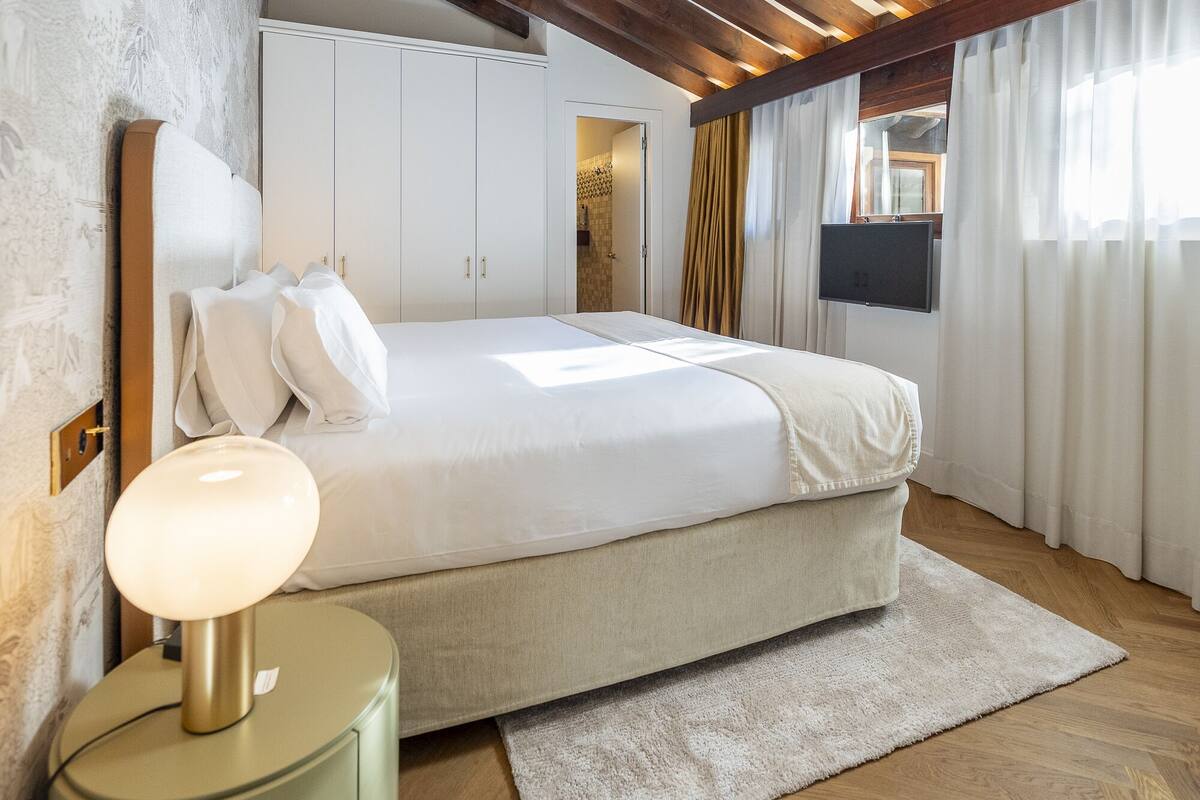 A bedroom with a large bed, a bedside lamp, wooden beams on the ceiling, and a TV mounted on the wall.