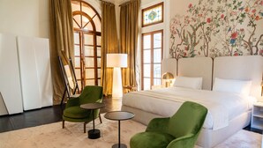 Quarto de hotel com uma cama grande, duas poltronas verdes, uma mesa redonda pequena e uma janela ampla com cortinas.