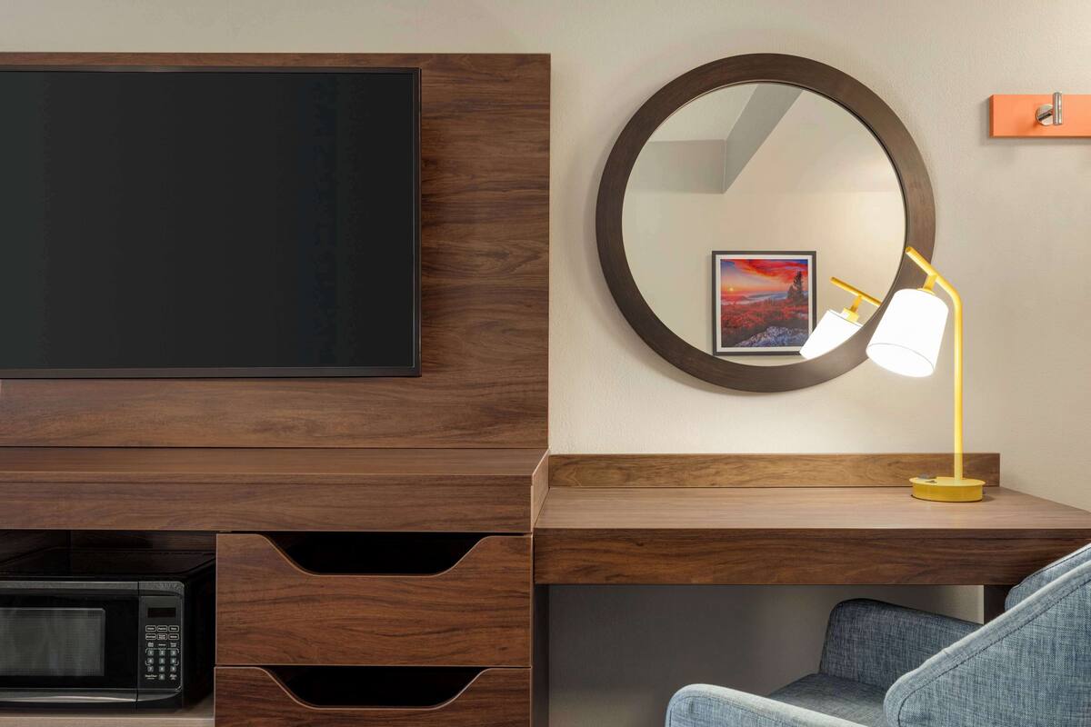 A modern hotel room with a flat-screen TV mounted on a wooden cabinet, a round mirror with a picture frame, a desk with a lamp, and a microwave.