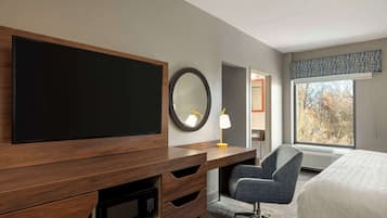 Ein Hotelzimmer mit einem großen Flachbildfernseher, einem Schreibtisch mit Schubladen, einem Sessel, einem Bett und einem Fenster mit Blick auf Bäume.