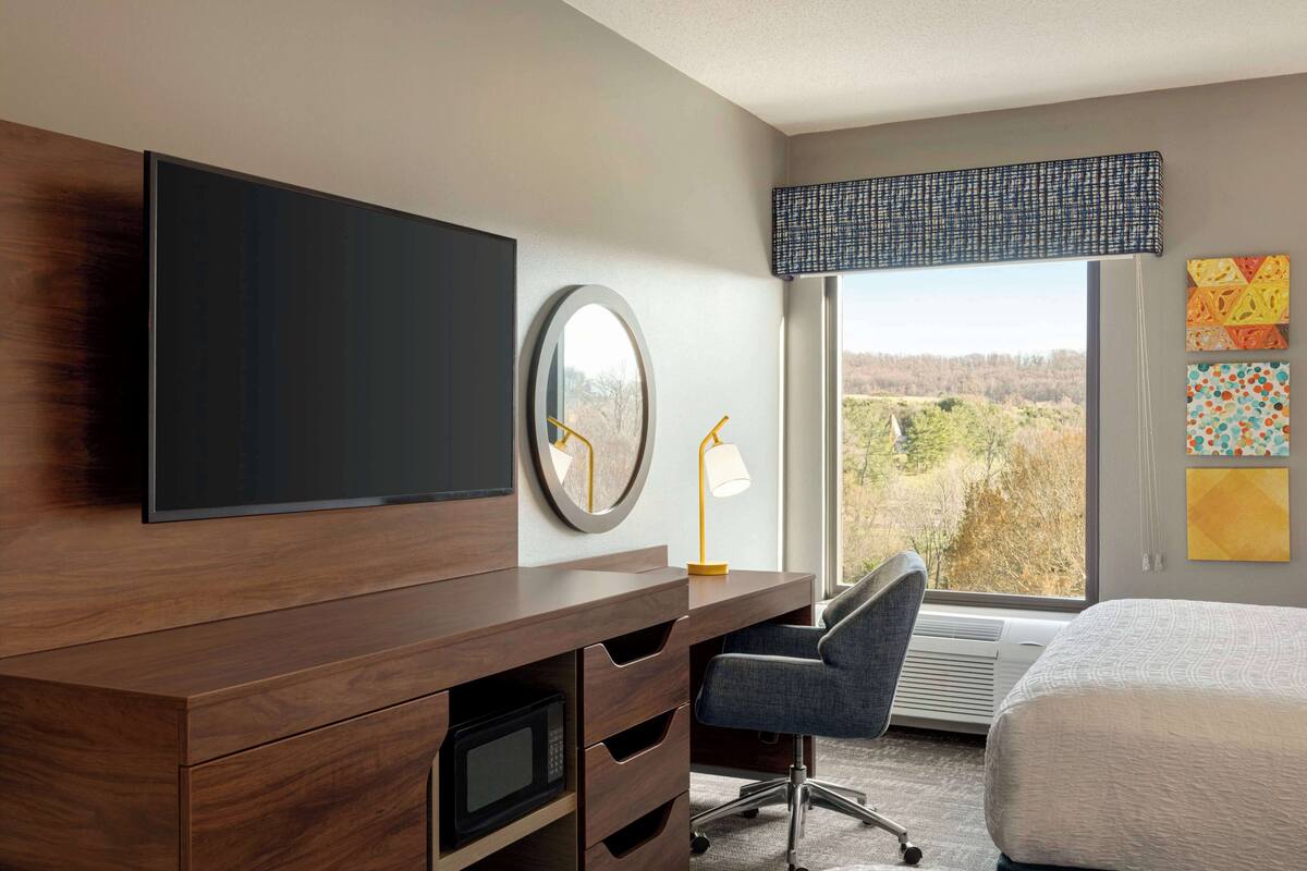 A hotel room with a wooden desk, a flat-screen TV, a round mirror, a desk lamp, a chair, a bed, and a large window with a view of trees.
