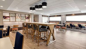 A modern dining area with wooden tables and chairs, a long communal table, and a ceiling with recessed lighting. - Hampton Inn Waynesboro/Stuarts Draft (Fishersville)
