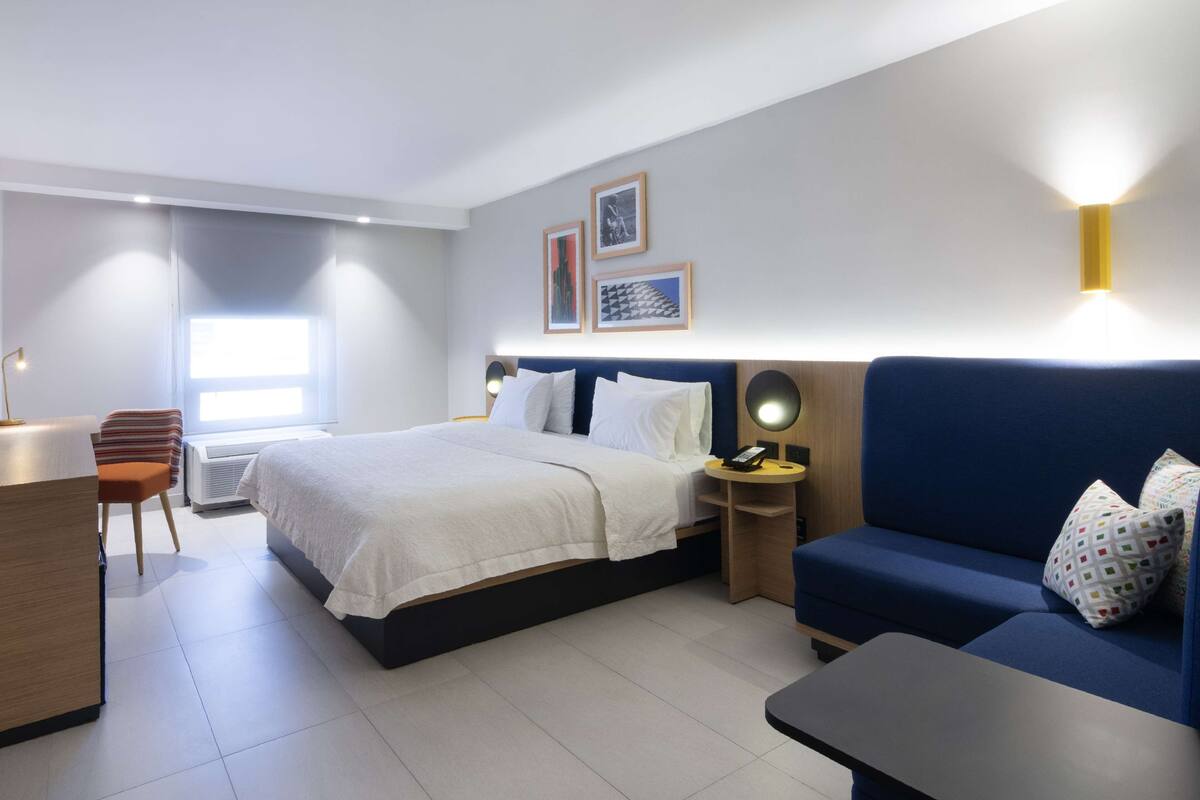 Ein modernes Hotelzimmer mit einem Bett, einem Schreibtisch, einem Sessel, einer Couch und einem kleinen Tisch. Zwei Nachttische mit Lampen und eine Wandleuchte sind vorhanden.
