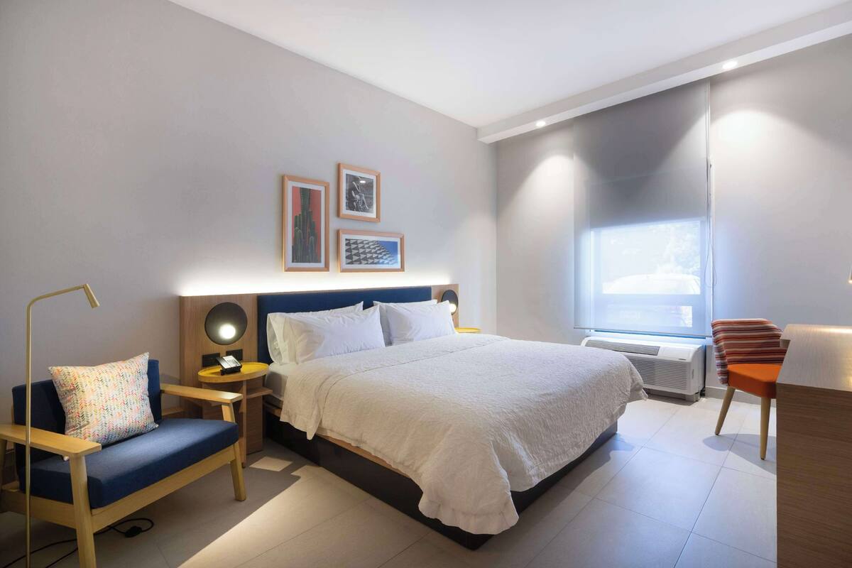 Ein modernes Hotelzimmer mit einem großen Bett, einem Schreibtisch, einem Stuhl und einem Nachttisch mit Lampe.