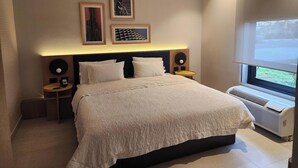 A modern bedroom with a large bed, a bedside table, a nightstand, a wall-mounted air conditioner, and framed artwork on the wall. - Hampton by Hilton Saltillo (Saltillo)