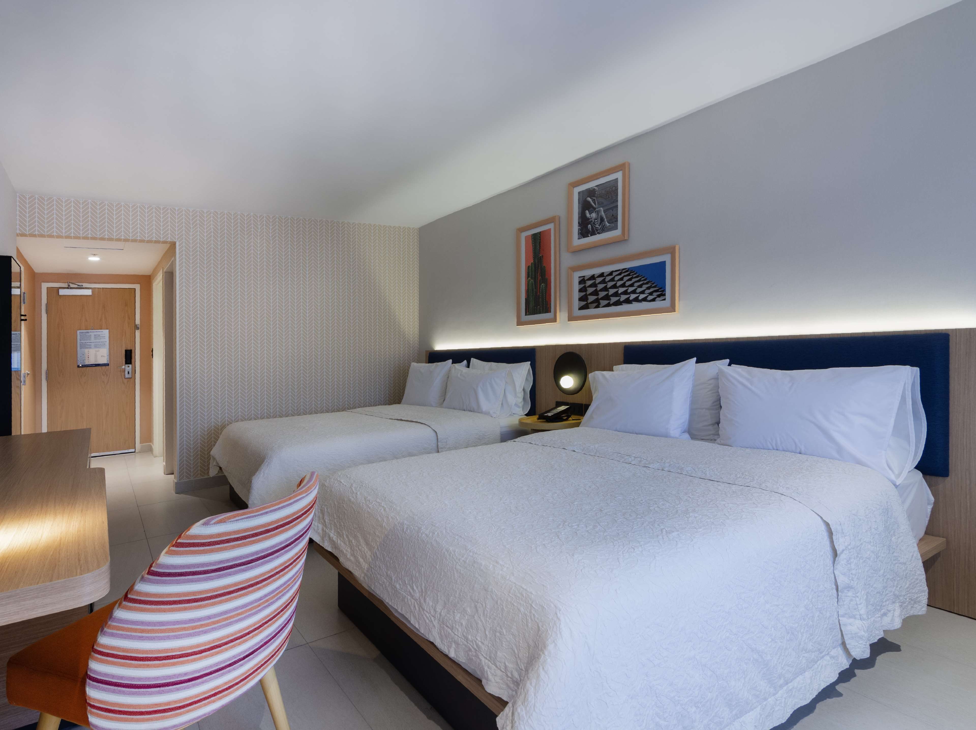 Habitación de hotel con dos camas, un escritorio y una silla. Se observan cuadros en la pared y una puerta con un cartel.