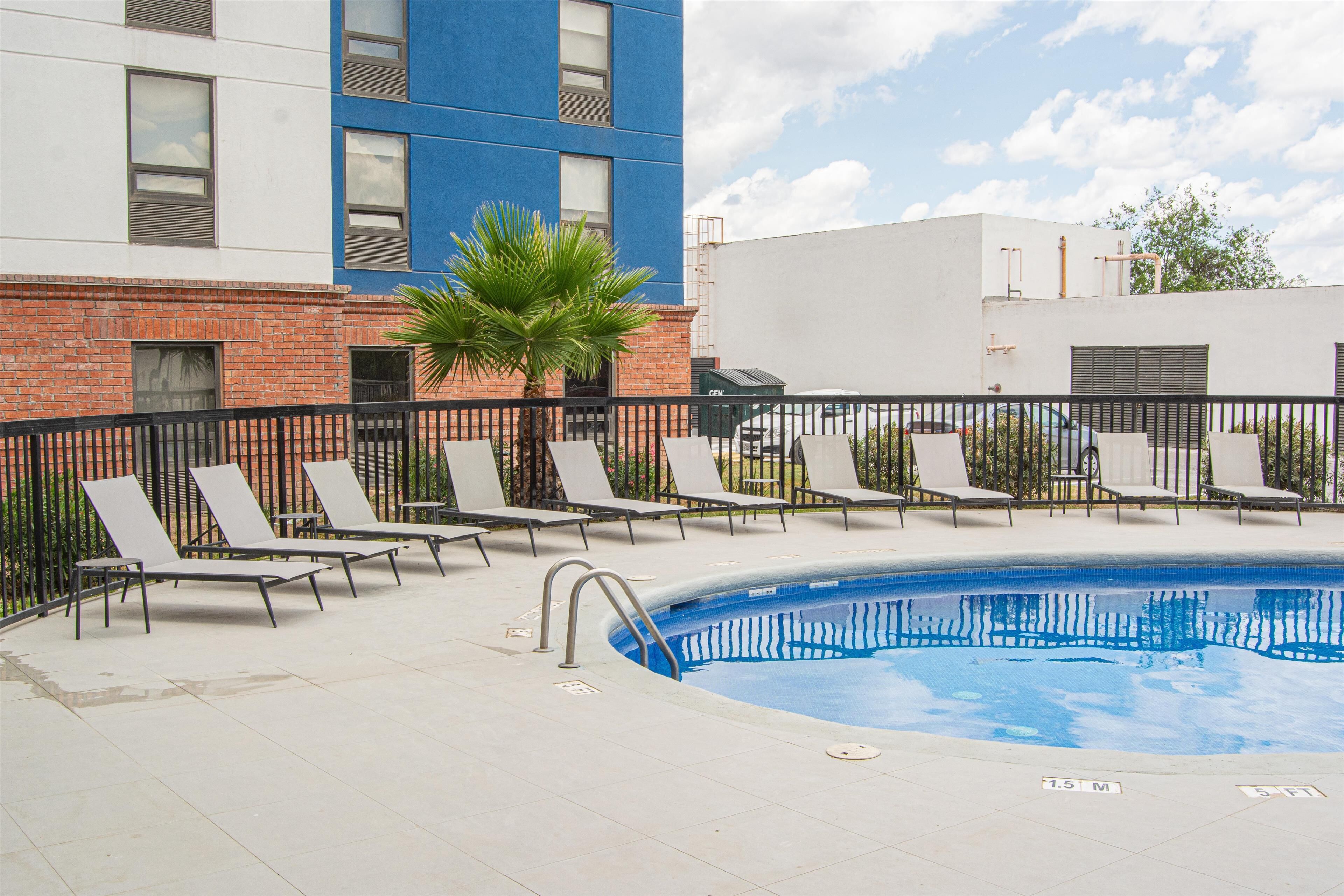 A swimming pool with lounge chairs and a palm tree, surrounded by a brick building and a white building.
