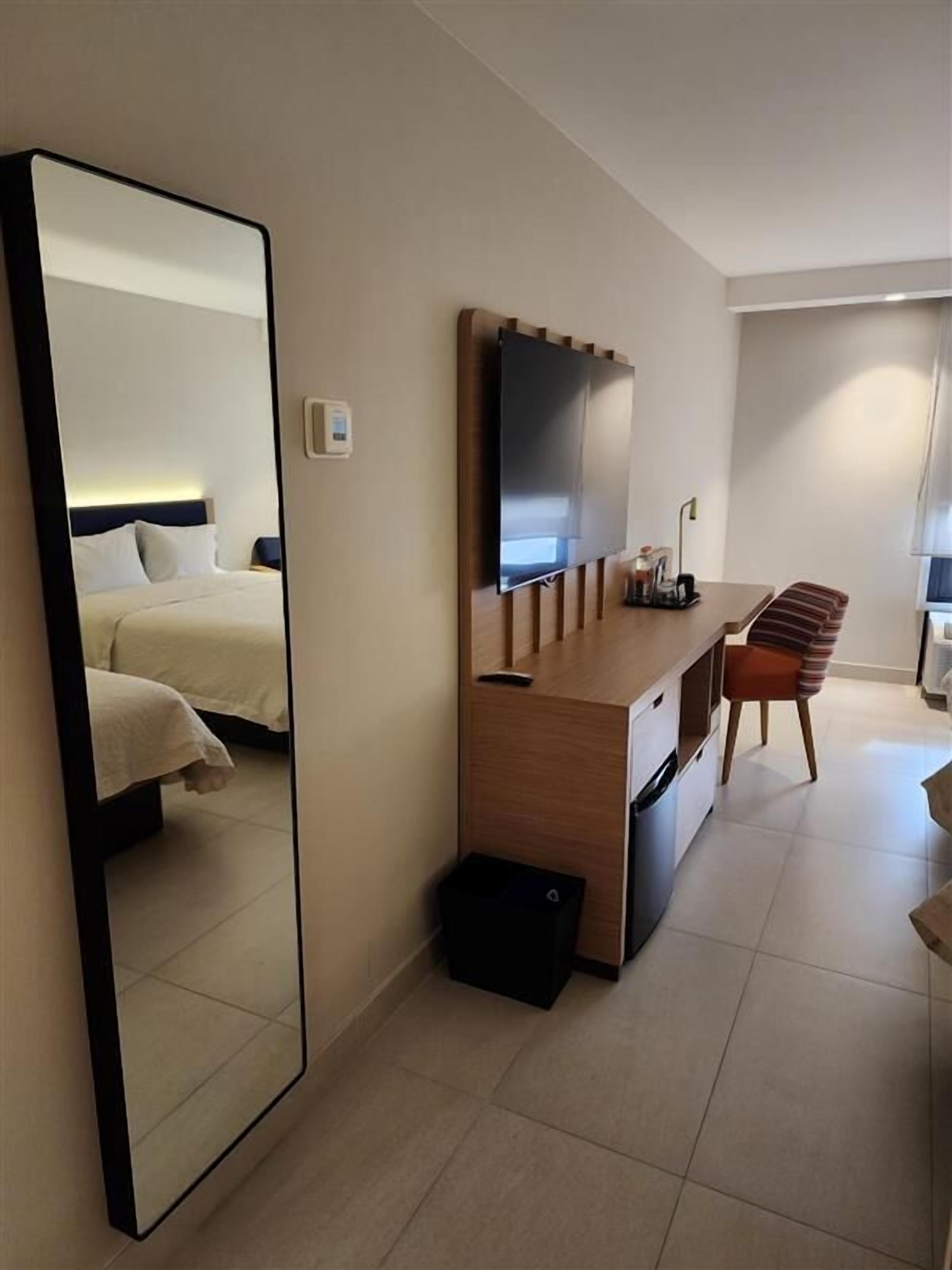 Ein Hotelzimmer mit einem Bett, einem Schreibtisch mit Stuhl, einem Fernseher und einem großen Spiegel.