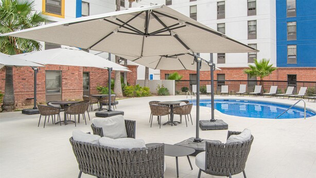 An outdoor seating area with wicker furniture and umbrellas, a pool, and a brick building in the background. - Hampton by Hilton Saltillo (Saltillo)