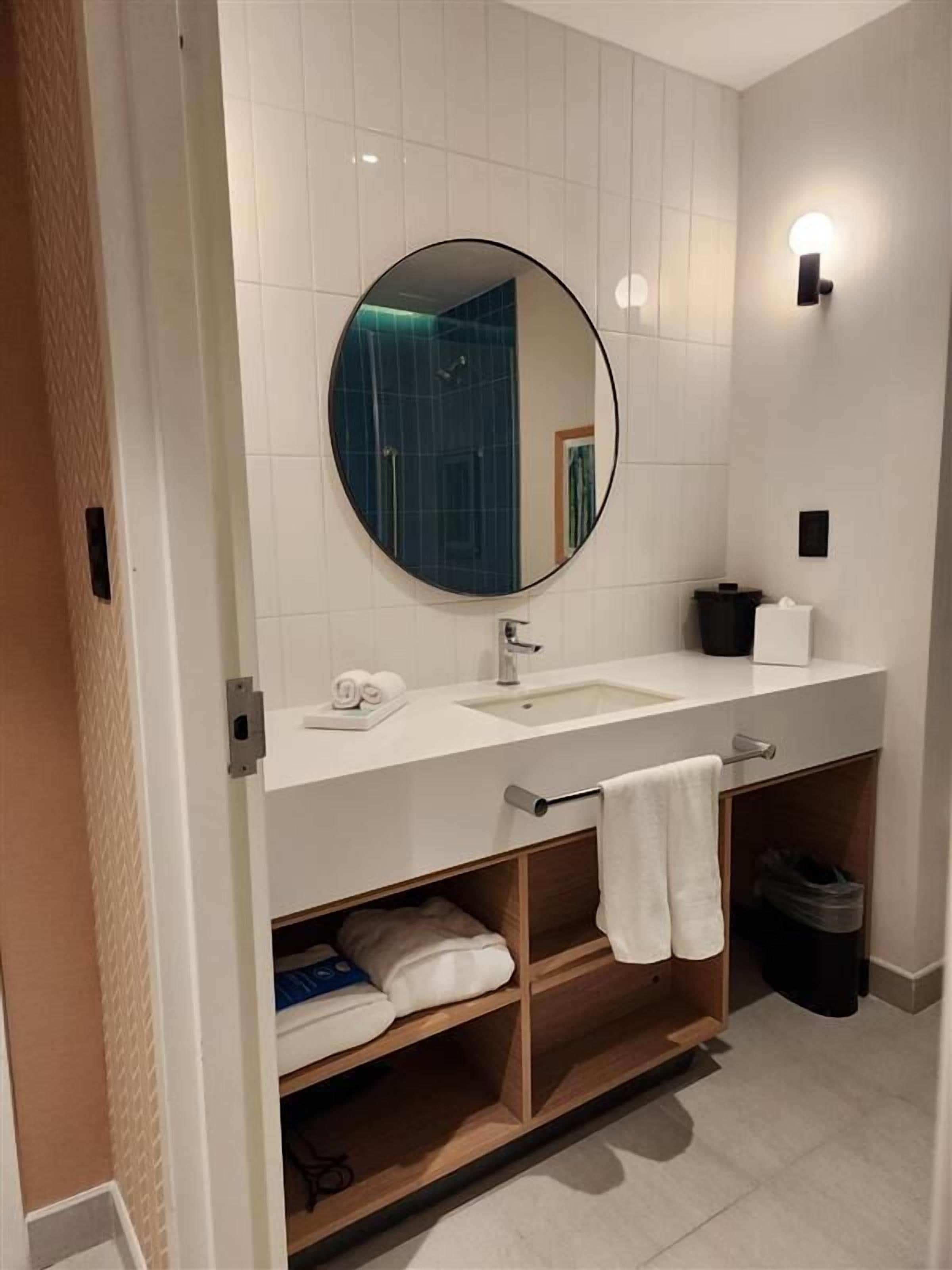 Un baño moderno con un tocador blanco, un espejo redondo, un perchero y una repisa con toallas dobladas.