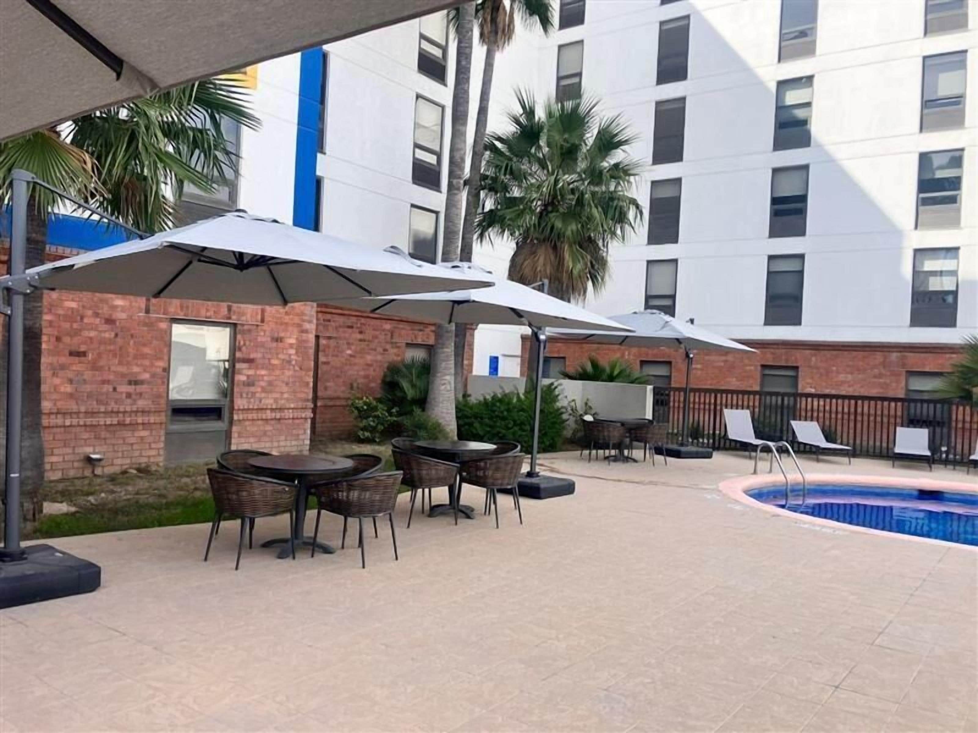 An outdoor patio area with tables and chairs under umbrellas, a pool, and a building with multiple windows in the background.