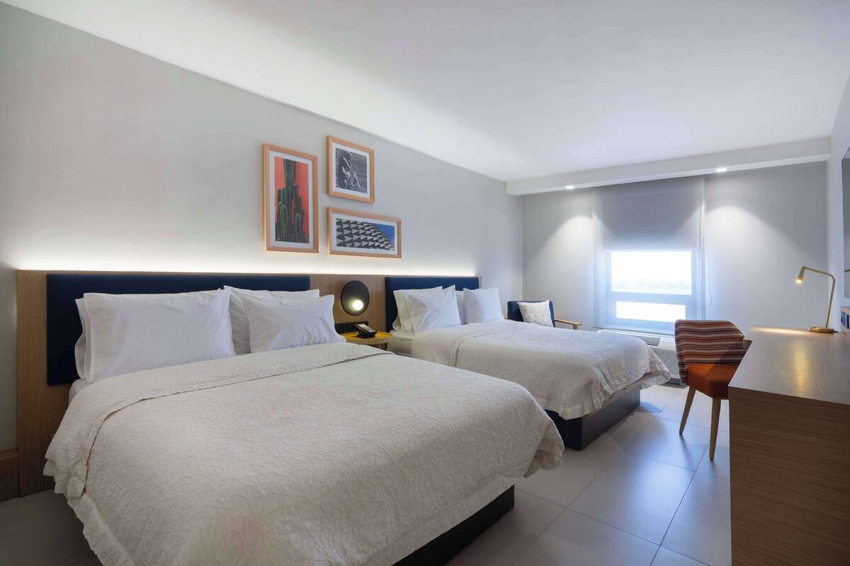 Ein Hotelzimmer mit zwei Betten, einem Schreibtisch und einem Stuhl. An der Wand befinden sich gerahmte Bilder, ein Fenster und eine Lampe auf dem Schreibtisch.