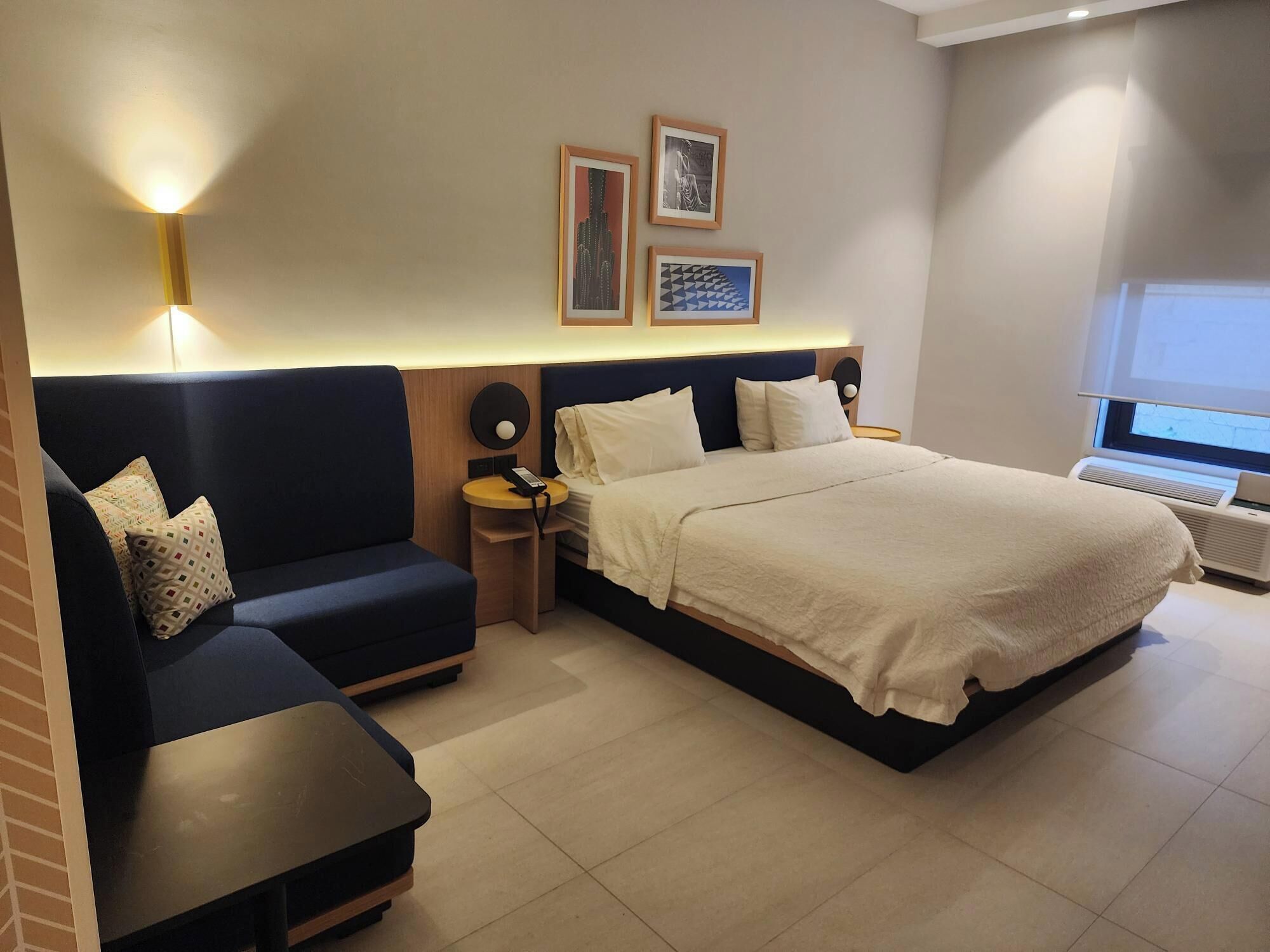A hotel room with a bed, a sofa, a small table, and a heater. There are framed pictures on the wall and a window with a view.