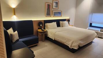 A hotel room with a bed, a sofa, a small table, and a heater. There are framed pictures on the wall and a window with a view.