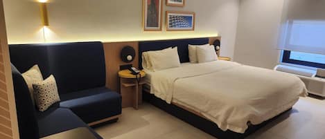 A hotel room with a bed, a sofa, a small table, and a heater. There are framed pictures on the wall and a window with a view.