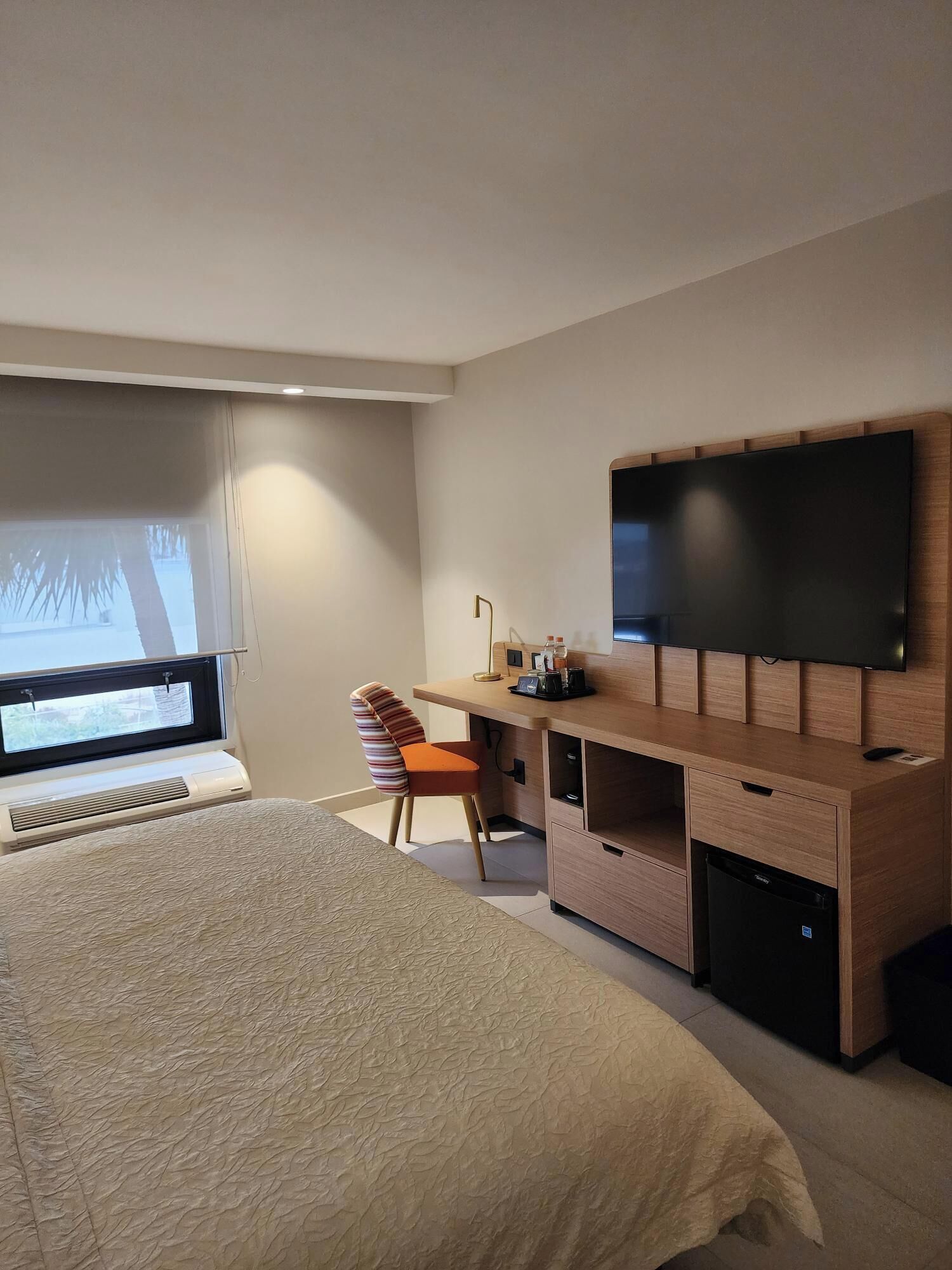 Una habitación de hotel moderna con una cama grande, un escritorio con televisor de pantalla plana, una silla y una ventana con persianas.