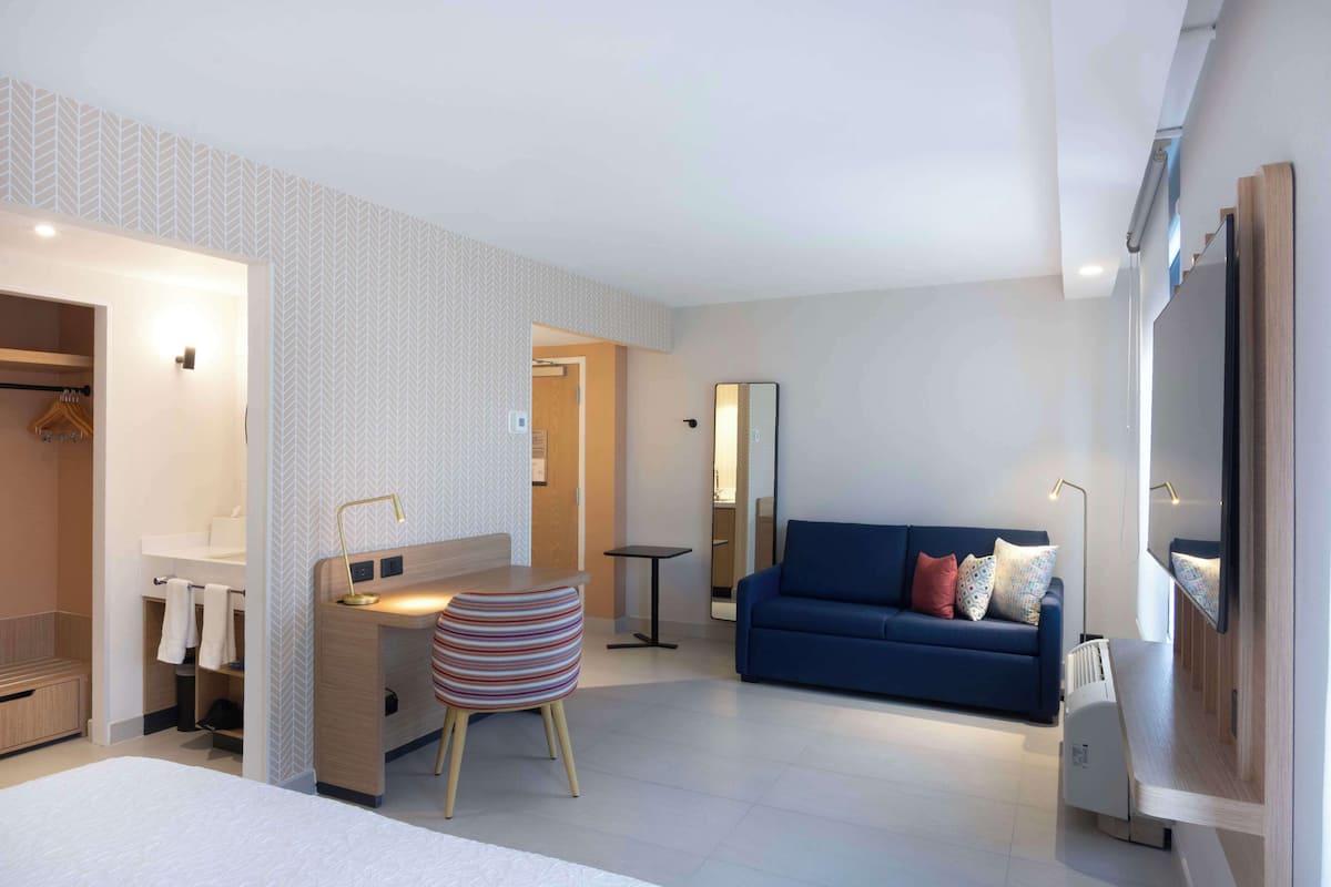 Ein modernes Hotelzimmer mit einem Bett, einem Schreibtisch mit Stuhl, einem blauen Sofa und einem Spiegel.
