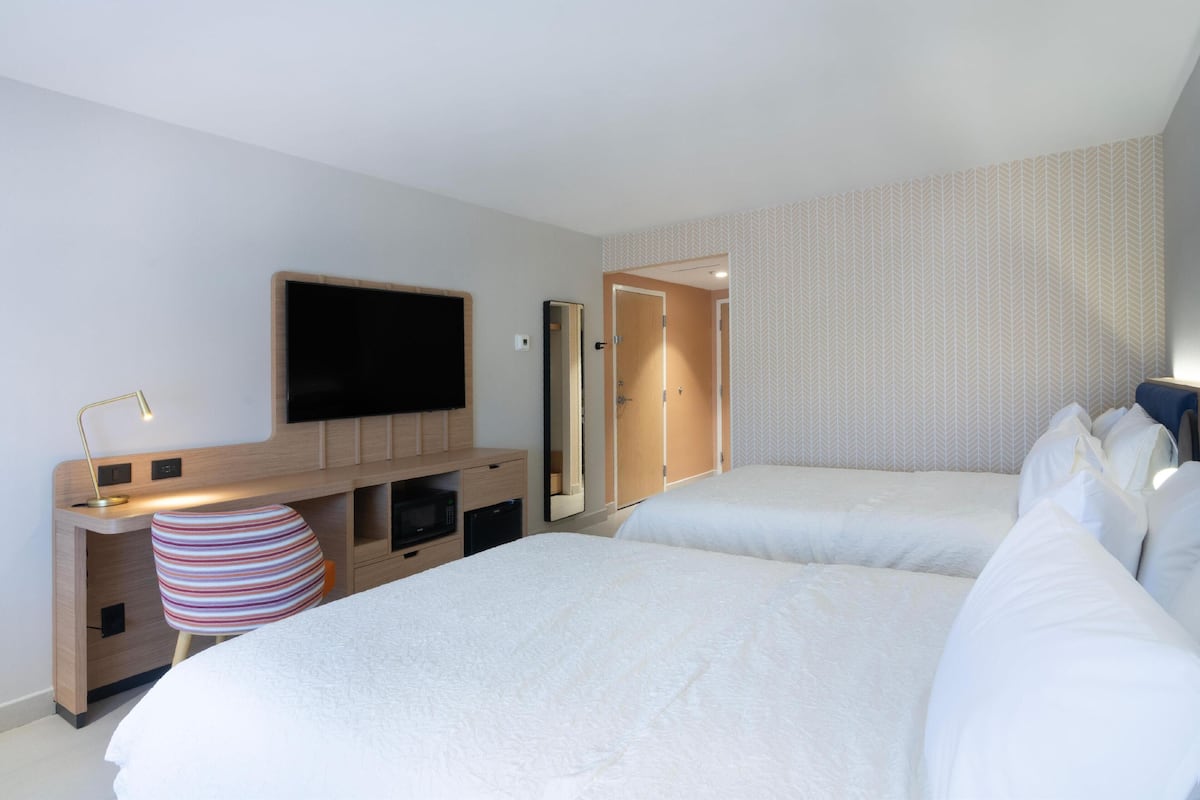 Ein Hotelzimmer mit zwei Betten, einem Fernseher, einem Schreibtisch mit Lampe und einem gestreiften Sessel.